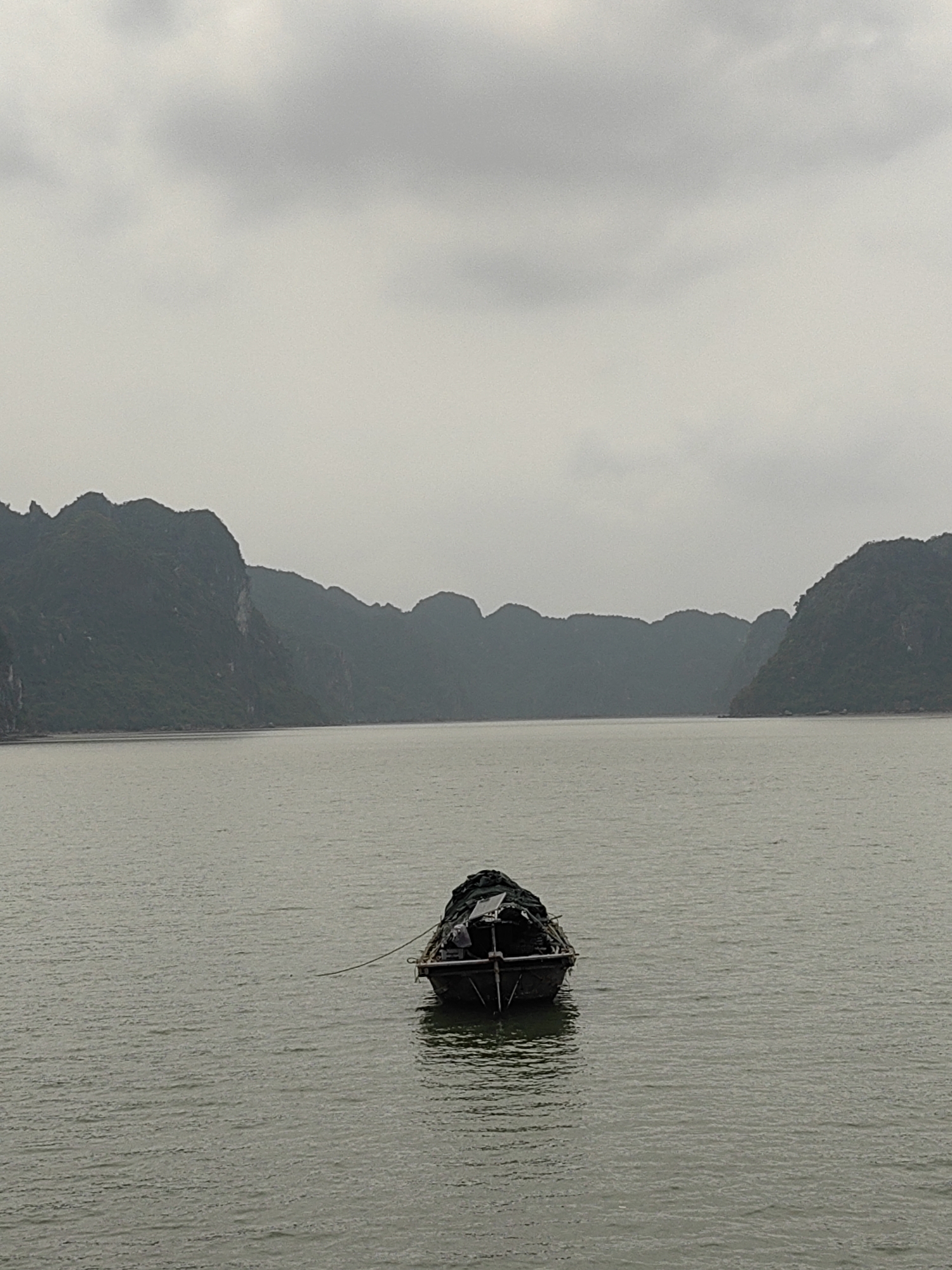 The image shows a small, dark-colored boat floating on water, with landmasses in the background. The sky is overcast, creating a muted, grey light. The boat is the central subject, and the background landmasses appear to be islands or mountains. The location is likely Ha Long Bay, in the city of Hải Phòng, Vietnam, given the geographic features. There are no people visible in the image. The water is relatively calm.
