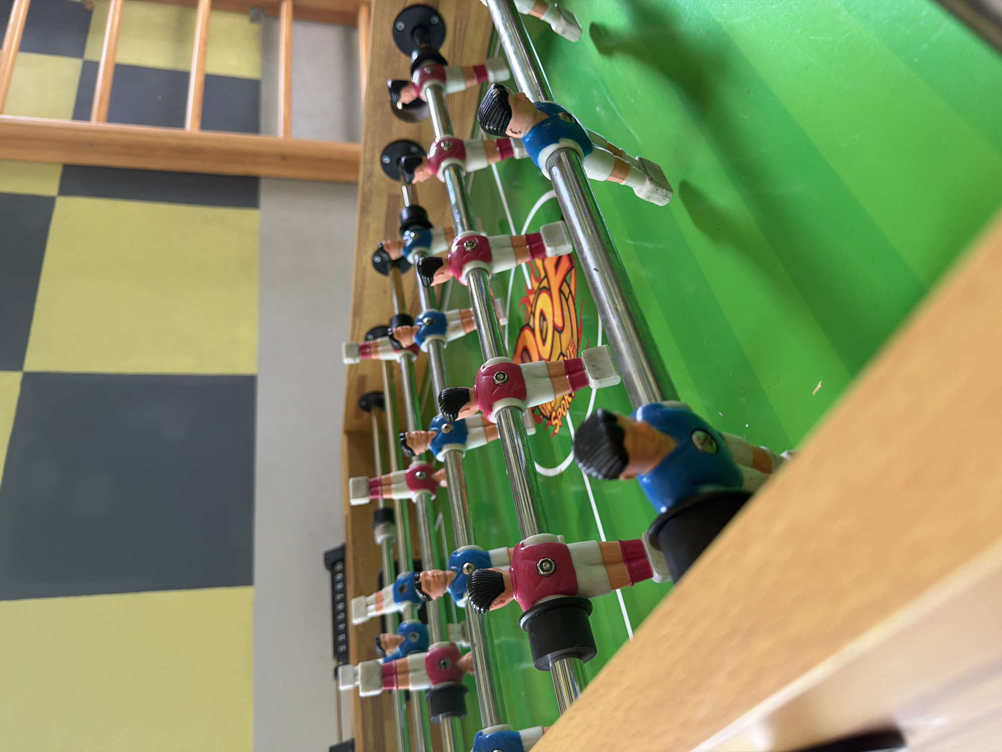 The image depicts a foosball table with blue and red teams, viewed from a low angle. The table is green with white markings. A wooden frame surrounds the playing field. To the left, the floor is tiled with yellow and gray squares. A partially visible wooden ladder-like structure is also present on the left. There is no indication of the location being Birobidzhan, Russia, or any other location. No people, actions, or interactions are visible.