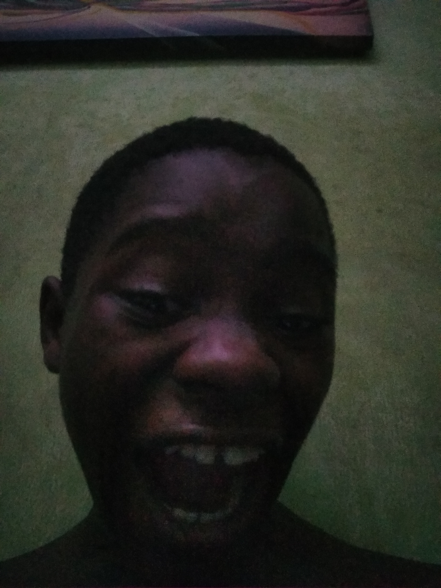 An image of a person with dark skin tone, cropped closely to the face. The person has short hair and is smiling widely, revealing their teeth. The background appears to be a dimly lit green wall, with a framed piece of artwork partially visible at the top of the image. The location is suggested to be Shangisha, Nigeria, but this is not directly evident from the image itself.