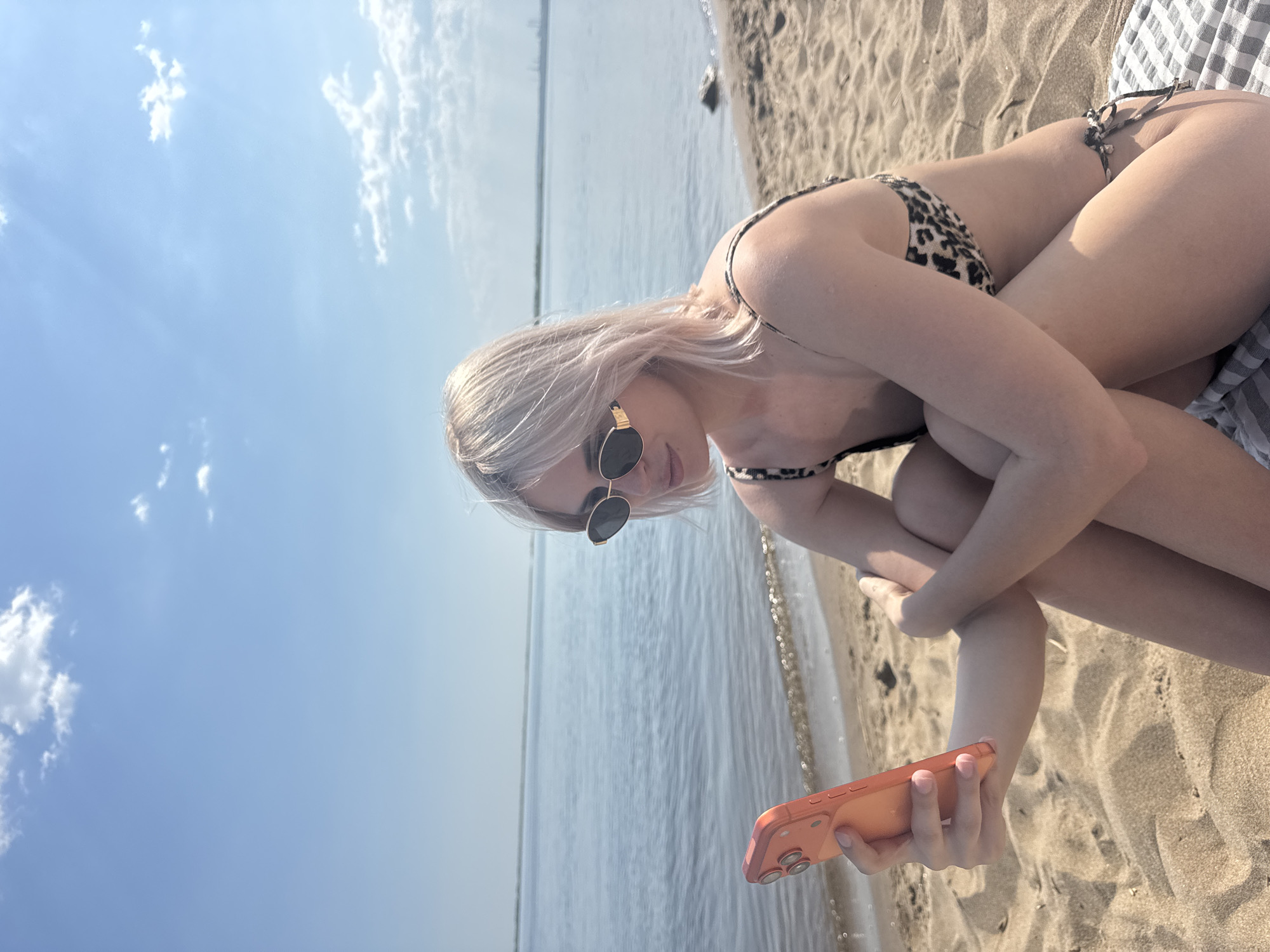 A woman is sitting on a beach next to a body of water. She is wearing a spotted bikini and sunglasses, and she is holding a phone in her hand. The beach is sandy, and there is a towel underneath her. The sky is blue with some clouds. The location is likely a beach in Saratov, Russia, given the request.