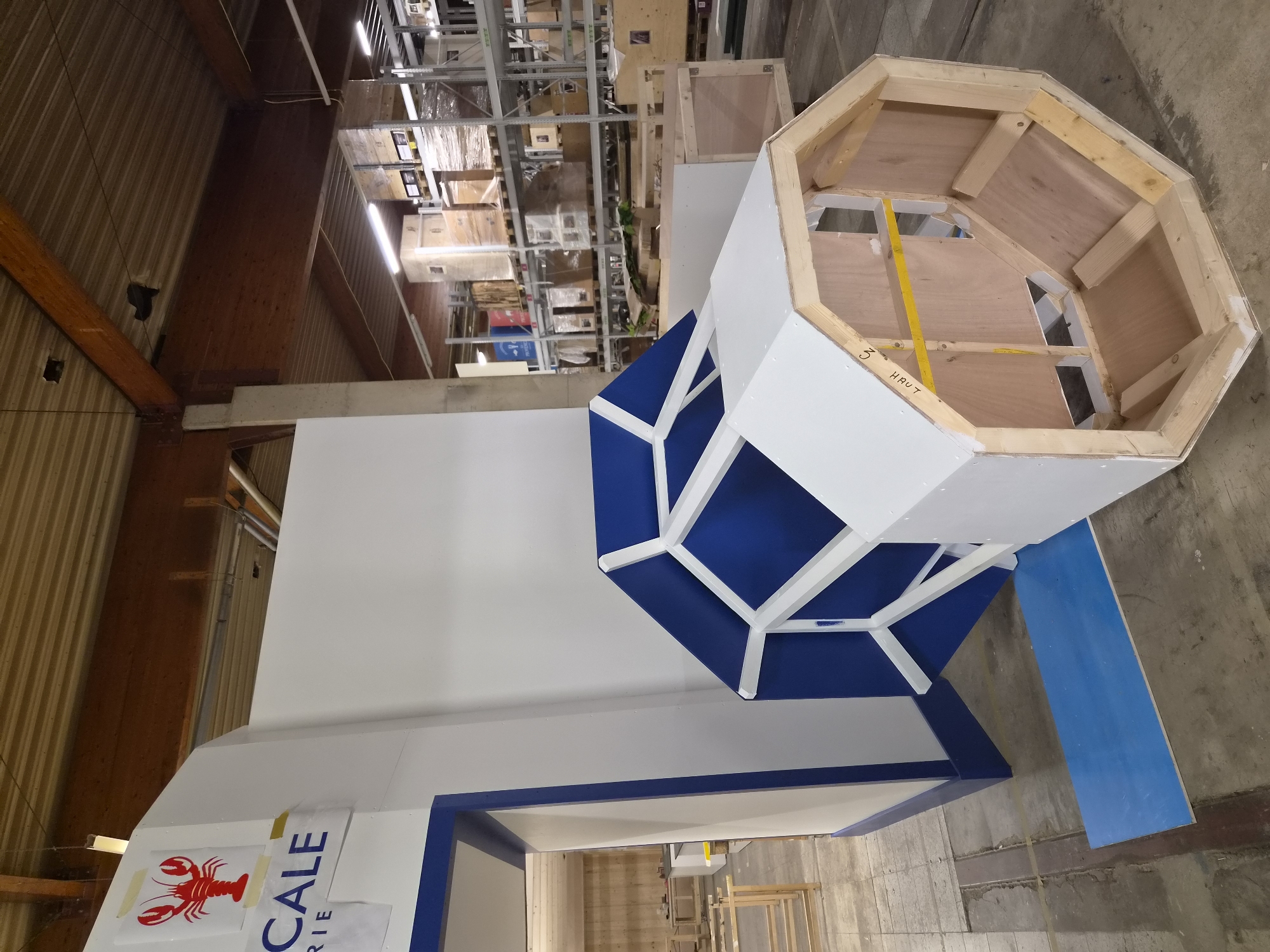 The image captures an indoor scene, likely a workshop or storage facility, located in Leudelange, Luxembourg. The foreground is dominated by a large, octagonal structure constructed with wood framing and white panels, with blue panels visible on the interior surfaces. This structure appears to be a part of a larger, custom-built installation, possibly for an exhibition or display. To the left, a portion of a white wall is visible, featuring a blue-trimmed doorway and a sign with a red lobster graphic and blue text that reads "CALE" and "RIE". The background reveals shelves stacked with various boxes and crates, suggesting a commercial or industrial environment. The floor is concrete, with some sections marked with tape. No people are visible in the image. The overall impression is of a construction or assembly process in progress within a warehouse-like setting.