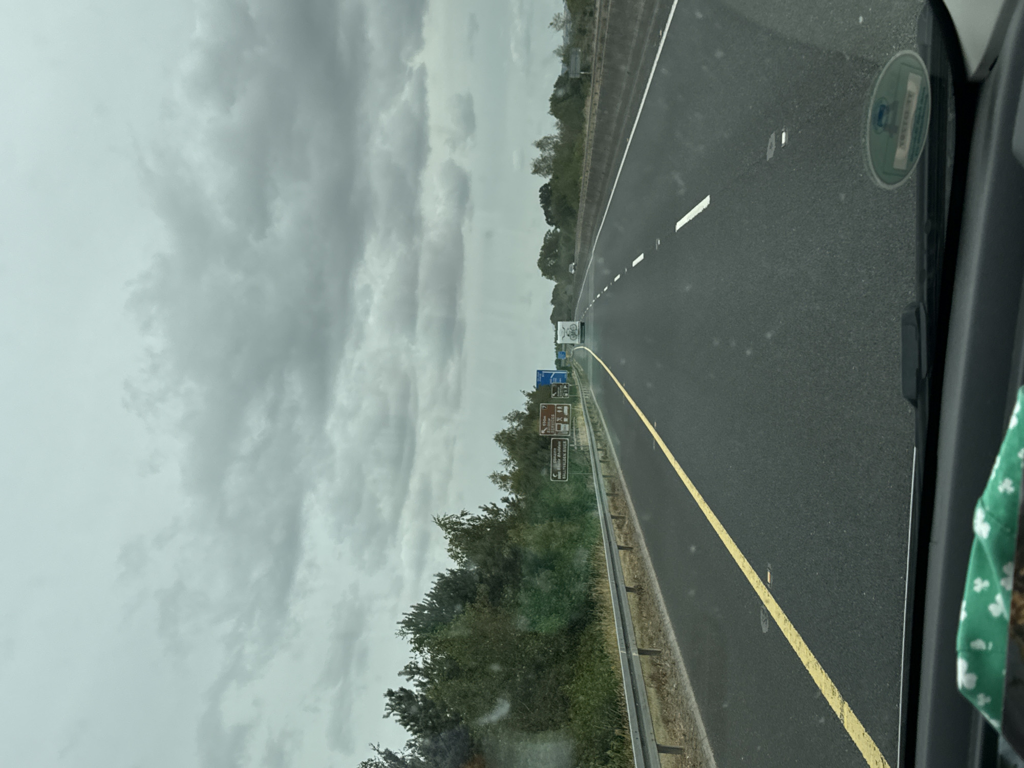 The image depicts a view from inside a vehicle traveling on a highway in Garryduff Lane, Ireland. The sky is overcast with dark clouds. On the left side of the highway, there is a row of trees and vegetation bordering the road. Several brown road signs are visible along the roadside. On the right side of the highway, there is a concrete barrier and more trees in the distance. The road itself is asphalt with a yellow line marking the left edge and dashed white lines indicating lanes. A white caravan with a bicycle graphic on its back is visible in the distance on the highway. The dashboard of the vehicle is partially visible at the bottom of the frame, with a green cloth adorned with shamrocks resting on it. There are also some small, reflective markers on the road surface.