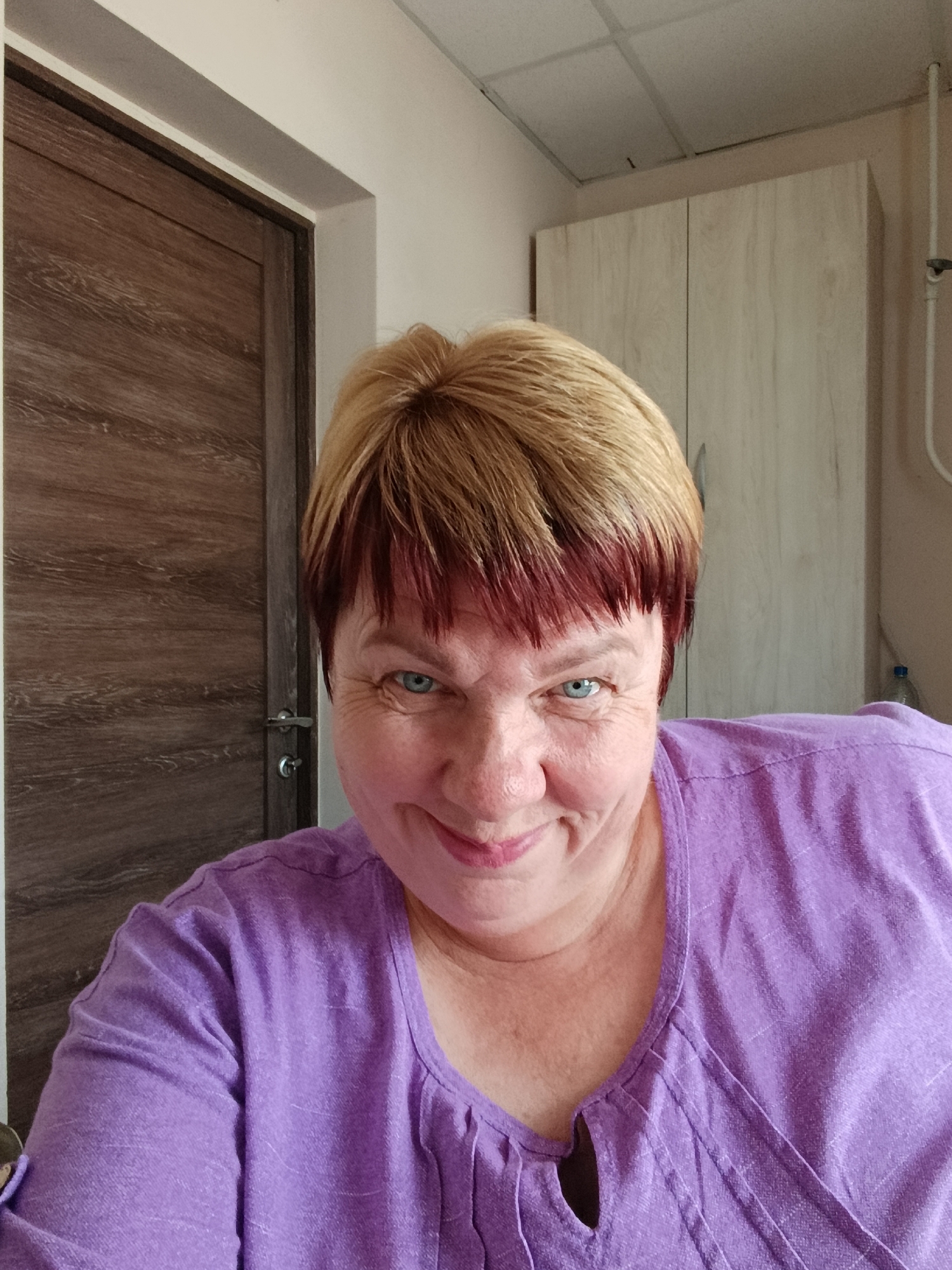 An adult woman with blonde hair with red undertones is taking a selfie indoors. She is wearing a purple shirt. The background includes a wooden door and a light-colored cabinet. The woman is smiling.