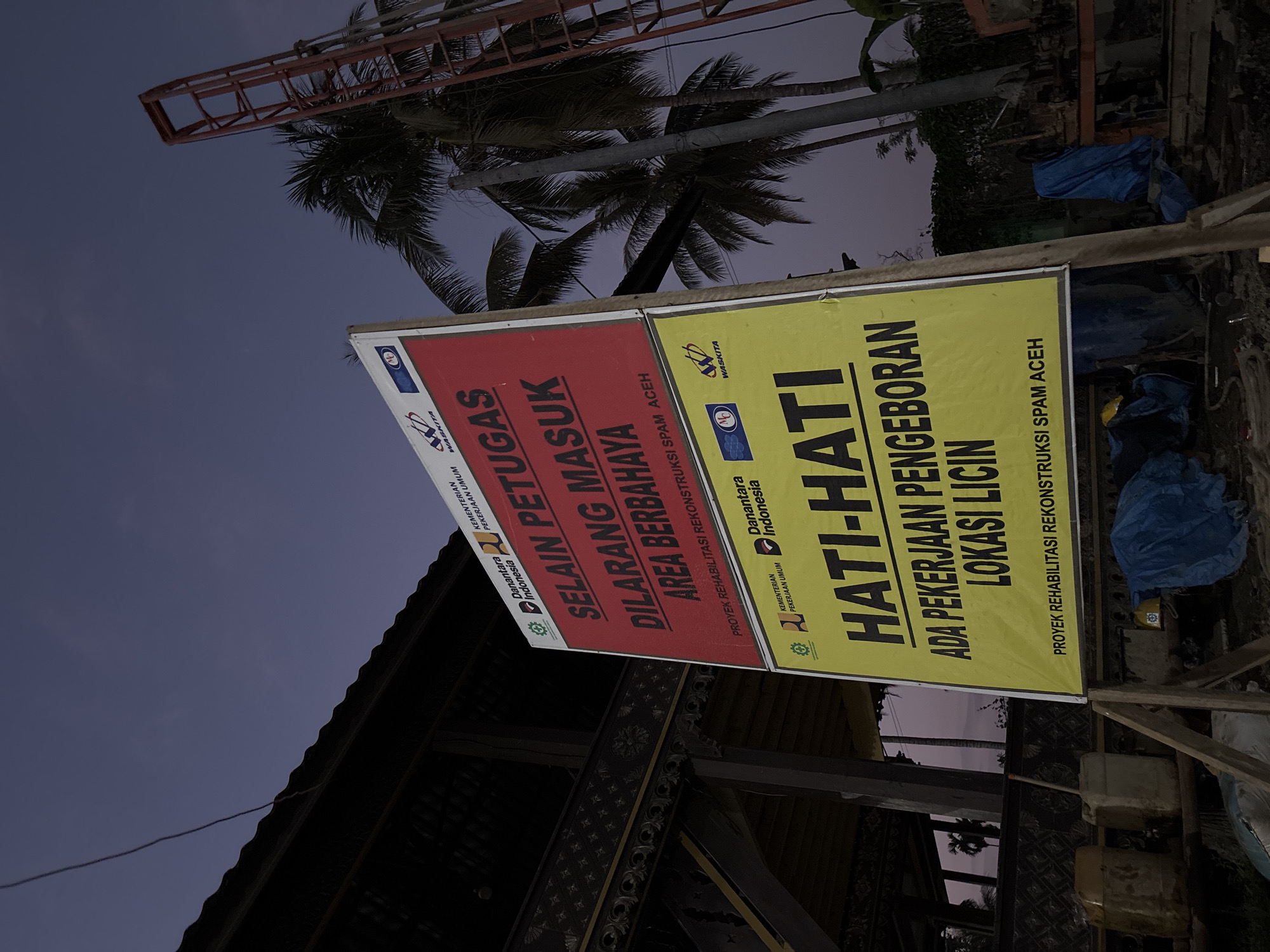 This media file captures an outdoor scene at what appears to be a construction or rehabilitation site in Mideuenjok, Indonesia, during dusk or early night, given the dark blue and purple hues of the sky. No people are visible.
The primary subjects are two large informational signs, mounted side-by-side on wooden posts. The left sign is predominantly red with white text, while the right sign is yellow with black text. Both signs bear logos for "Danantara Indonesia," "KEMENTERIAN PEKERJAAN UMUM" (Ministry of Public Works), and "WASKITA" (a construction company).
The red sign states: "SELAIN PETUGAS DILARANG MASUK AREA BERBAHAYA," which translates to "EXCEPT FOR OFFICERS, ENTRY TO DANGEROUS AREA IS PROHIBITED." Below this, it identifies the activity as "PROYEK REHABILITASI REKONSTRUKSI SPAM ACEH," meaning "REHABILITATION AND RECONSTRUCTION PROJECT OF SPAM ACEH" (likely referring to a Drinking Water Supply System).
The yellow sign reads: "HATI-HATI," meaning "BE CAREFUL" or "CAUTION." It further warns: "ADA PEKERJAAN PENGEBORAN LOKASI LICIN," translating to "THERE IS DRILLING WORK, SLIPPERY LOCATION." This sign also identifies the project as "PROYEK REHABILITASI REKONSTRUKSI SPAM ACEH."
In the background, several palm trees are silhouetted against the dim sky. Parts of a building structure, possibly a traditional wooden building with intricate carvings, are visible beneath the signs. On the ground near the yellow sign, blue tarps and some containers can be seen, hinting at ongoing construction or storage of materials. The overall scene conveys a cautionary message about an active construction or infrastructure rehabilitation project in a tropical setting.