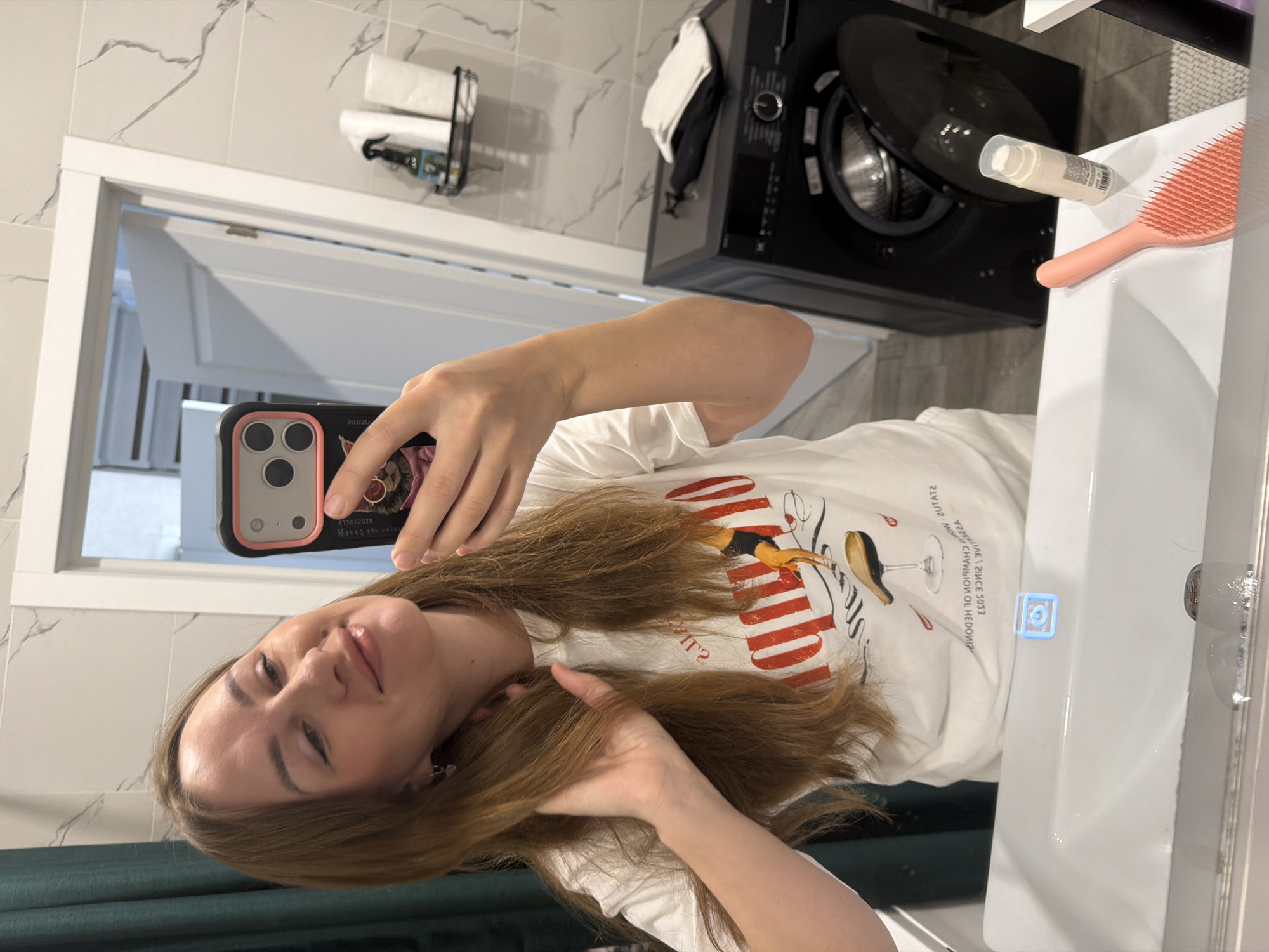 A woman is taking a mirror selfie in a bathroom. She is wearing a white t-shirt with a graphic design and has long, brown hair. She is holding her phone in her right hand and touching her hair with her left. In the background, there is a washing machine, a sink, a shelf with toiletries, and a closed door. The bathroom appears to be modern with tiled walls. There is no indication that this location is in Malinovka, Russia.