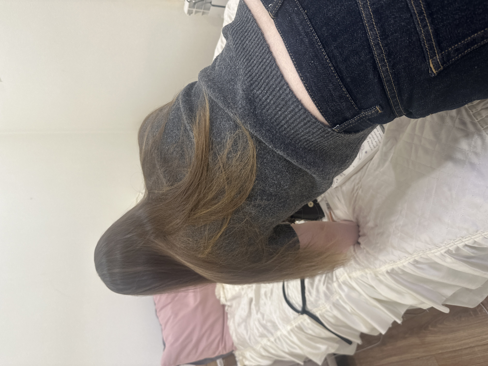 A person with long brown hair is lying on a bed. They are wearing a gray sweatshirt and dark blue jeans. The bed has a white quilted comforter with a ruffled edge. A pink pillow is visible beneath the person's head. The background is a plain white wall. The location is unknown and cannot be determined from the image.
