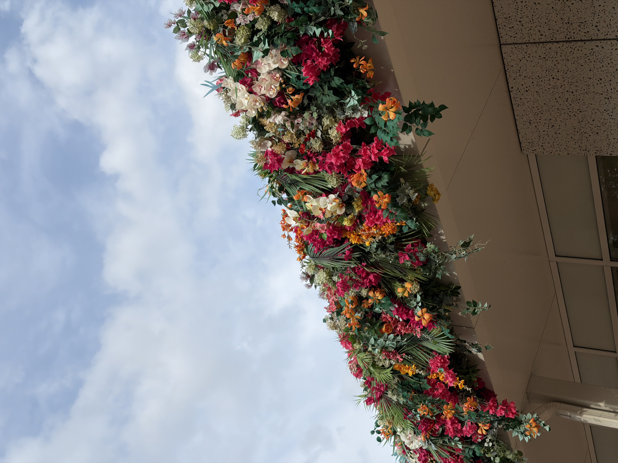 The image shows a dense arrangement of artificial flowers and greenery cascading over the edge of a building's overhang. The flowers are vibrant and varied in color, including shades of pink, red, orange, and white, with numerous green leaves and fronds interspersed throughout. The overhang appears to be made of a light-colored material, possibly concrete or painted wood. Below the overhang, the upper portion of a building facade is visible, featuring large windows and speckled textured panels. The sky above is a pale blue with scattered white clouds. There is no indication of the specific location in Moscow, Russia.