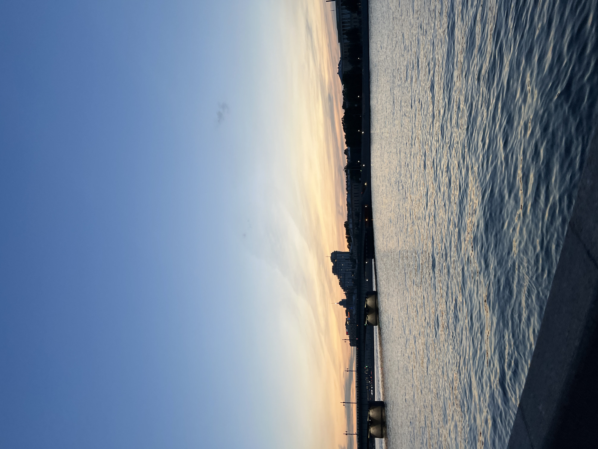The image depicts a serene river scene at dusk in an unidentified city in Russia. A wide river flows in the foreground, with gentle ripples on its surface reflecting the soft hues of the sky. In the mid-ground, a bridge spans the river, with its illuminated supports casting a warm glow on the water. Beyond the bridge, the city skyline is visible, characterized by a silhouette of buildings and trees against the fading daylight. The sky transitions from a deep blue at the top to lighter shades of orange and pink near the horizon, indicating the setting of the sun. A few wispy clouds are scattered across the sky, adding texture to the twilight atmosphere. The overall impression is one of peaceful urban twilight.