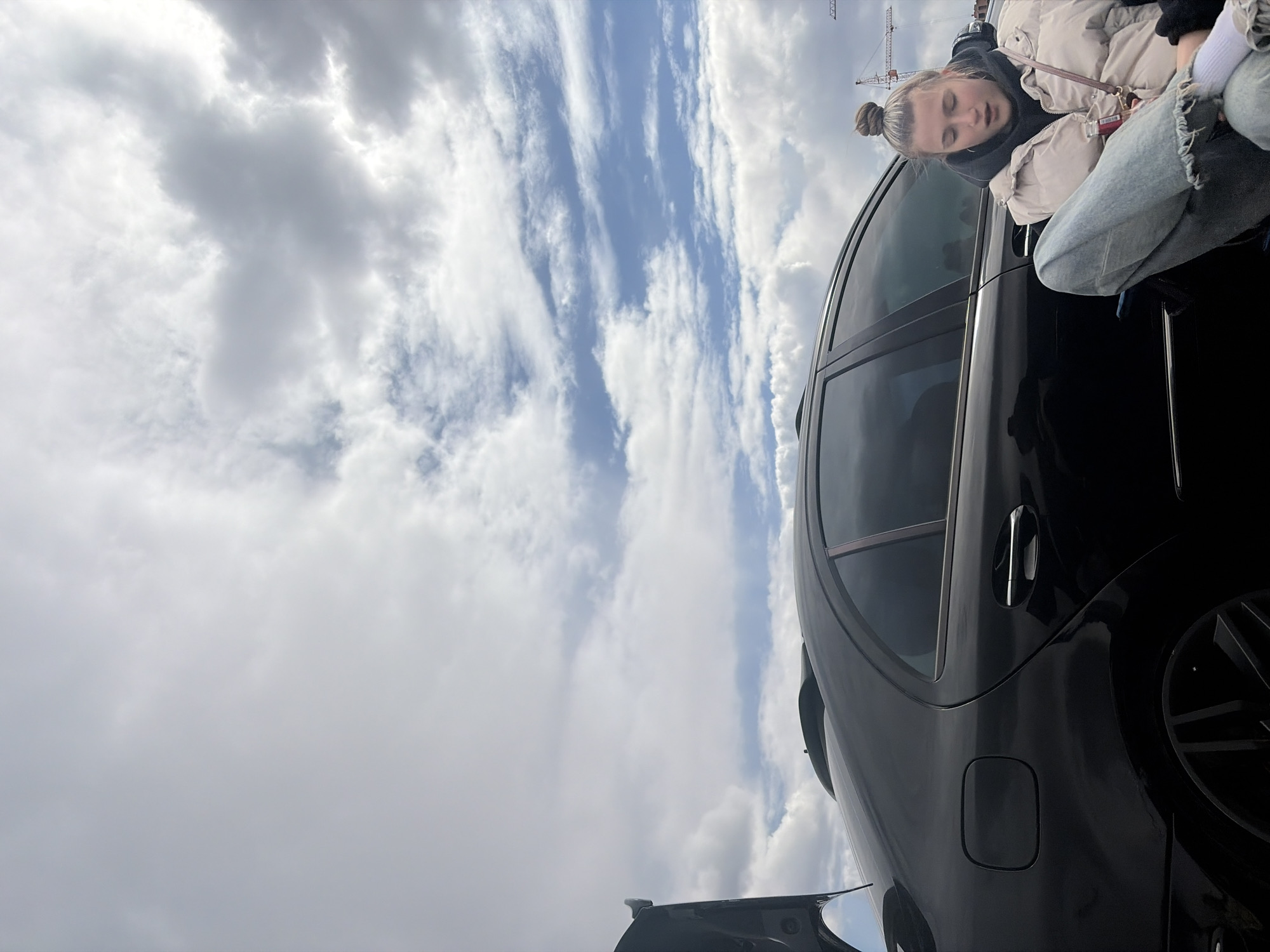 The image captures a young woman sitting on the roof of a black car. She is wearing a beige puffer jacket, a dark hoodie, and jeans. The background consists of a cloudy sky. The location is unidentifiable as Perm, Russia based on visual information alone.