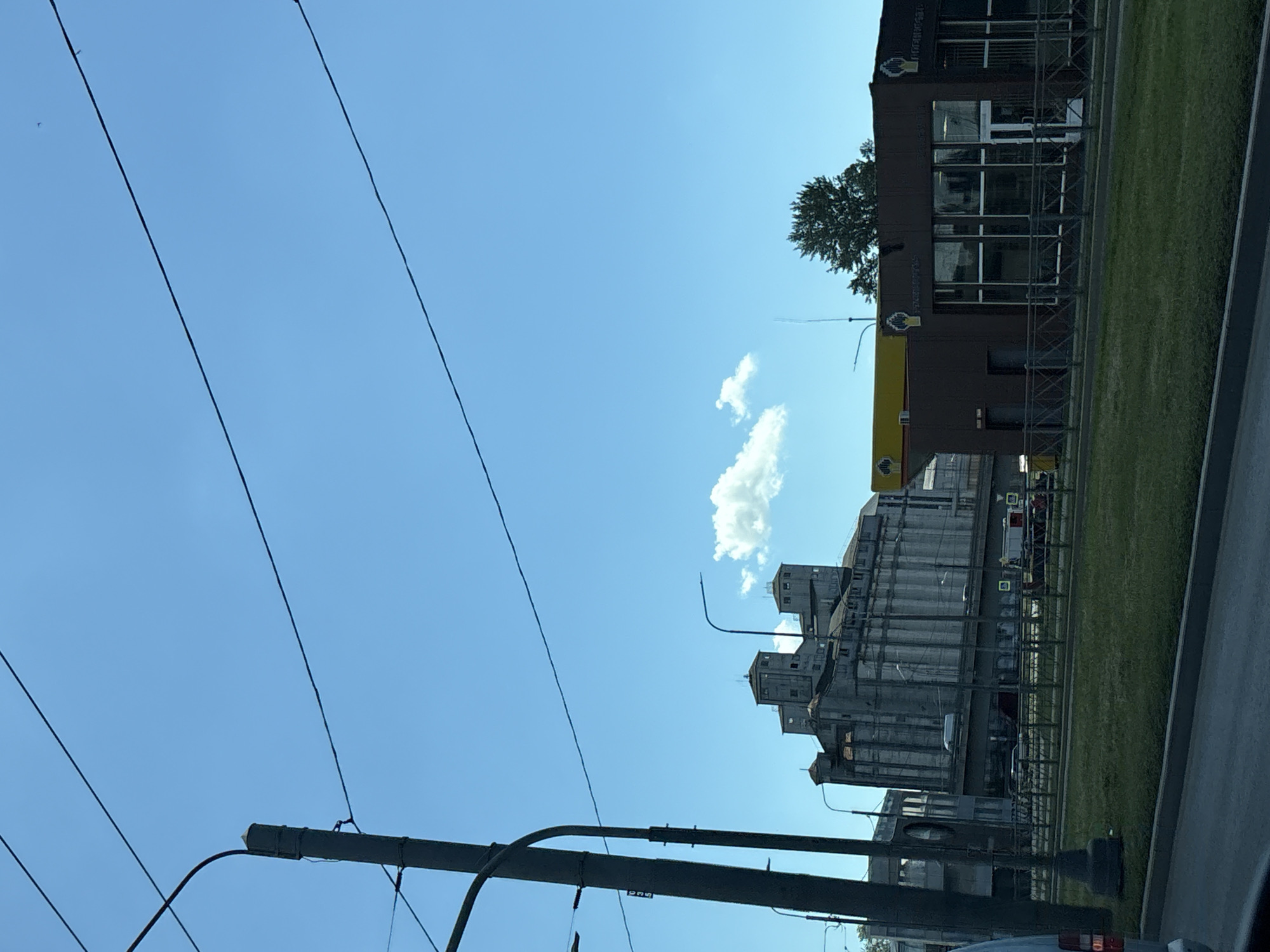 In Saint Petersburg, Russia, a large, multi-story industrial building with a tiered structure of silos stands under a clear blue sky. In the foreground, a dark, modern building with large glass windows and a yellow awning is visible. A chain-link fence separates the sidewalk from a patch of green grass. Overhead power lines crisscross the sky, and a solitary, fluffy white cloud drifts by. The scene appears to be a snapshot from a moving vehicle, suggesting a daytime street-level view.