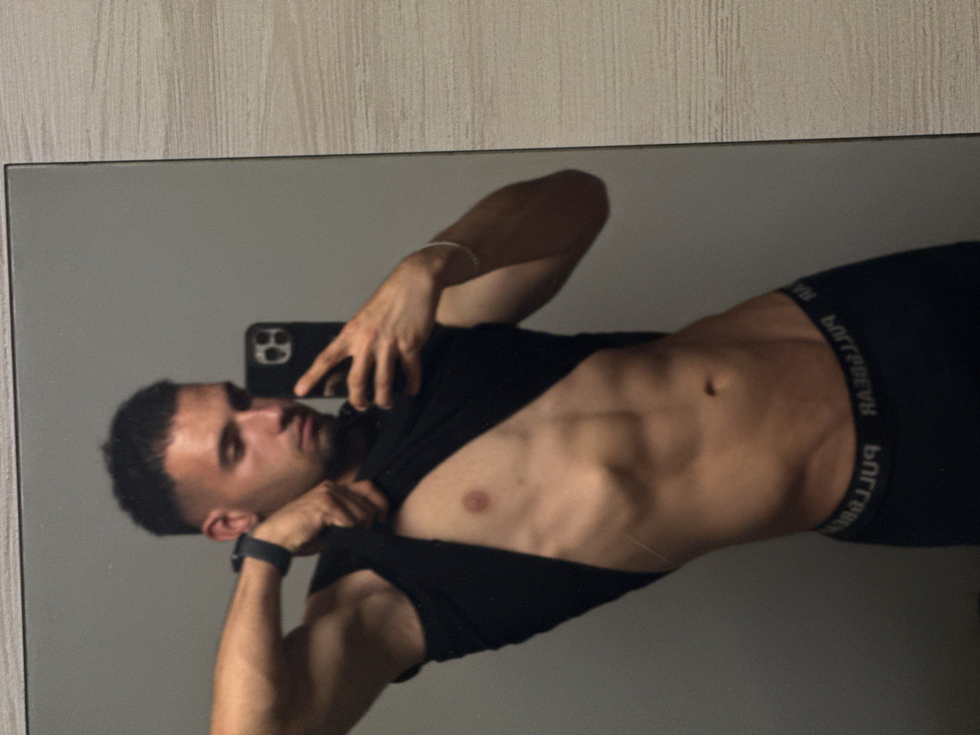 A young adult male is depicted in a mirror selfie, with the image rotated 90 degrees clockwise. He has short, dark hair and a muscular physique, evident in his arms, chest, and abdomen. He is holding a smartphone, identifiable as an iPhone model with a three-camera system, in his right hand to capture the photo. He wears a black t-shirt, which he has lifted to reveal his torso, and black boxer briefs or underwear with a white elastic waistband. A dark watch is visible on his left wrist, and a thin, light-colored bracelet adorns his right wrist.
The man's expression is neutral as he looks towards the phone's reflection. The setting appears to be indoors, with the reflection showing a plain, light gray background, likely a wall or surface behind him. Above the mirror's edge, a light-colored, subtly textured wooden wall is visible. The lighting is consistent with an indoor environment. On the elastic waistband of his underwear, the brand name "PULL&BEAR" is clearly visible in white, appearing mirrored as "RAEB&LLUP."