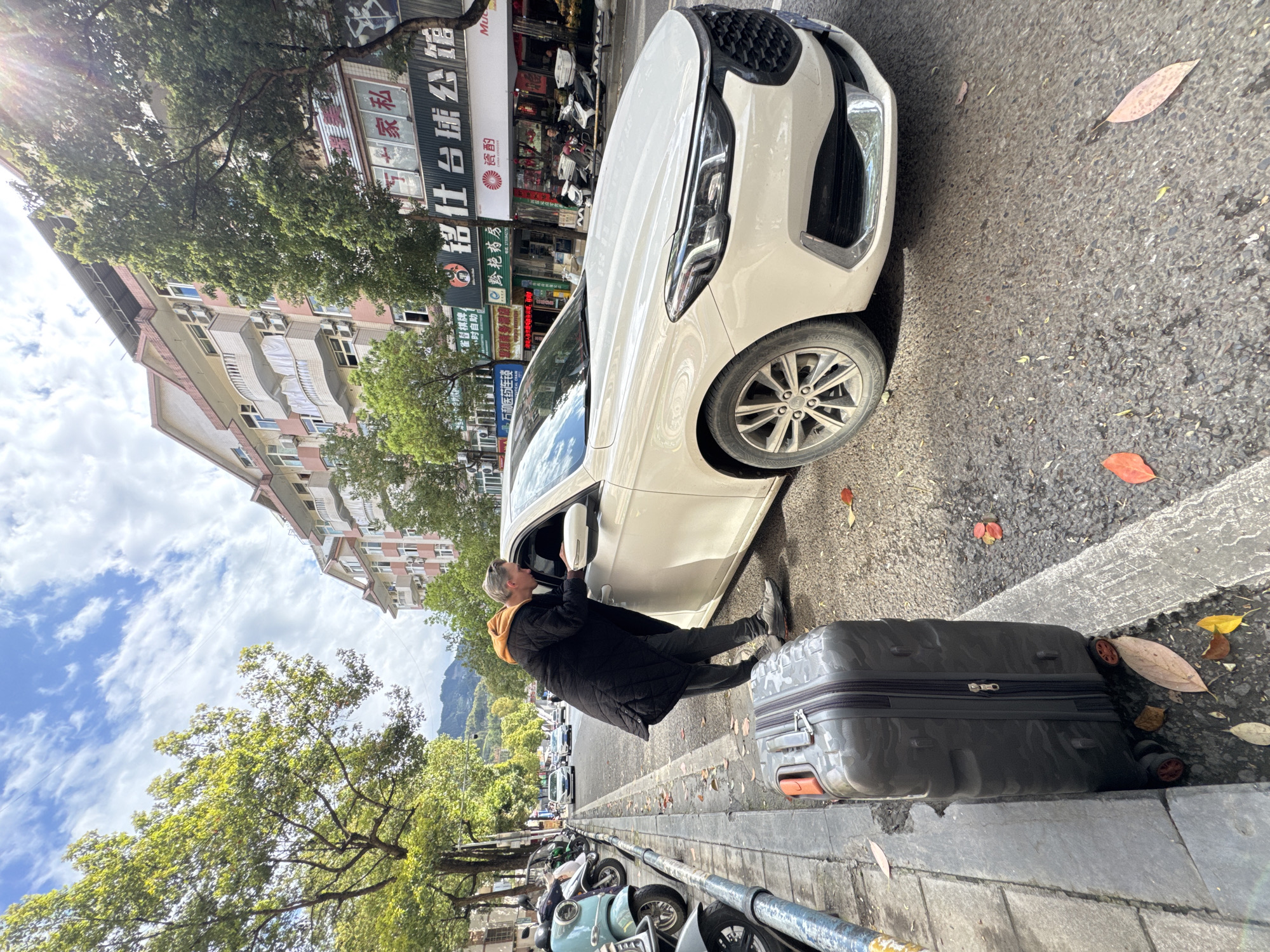 The image depicts a street scene in Qianjiang, China. A light-skinned person, dressed in dark clothing with a visible orange hoodie, is interacting with the driver-side mirror of a white sedan. A gray suitcase sits on the sidewalk next to the car. Parked scooters line the curb beyond the car. The background shows a building with Chinese signage and a mix of commercial and residential units, partially obscured by trees. The sky is visible with a mixture of blue and clouds. Fallen leaves are scattered on the ground, indicating an autumn season.
