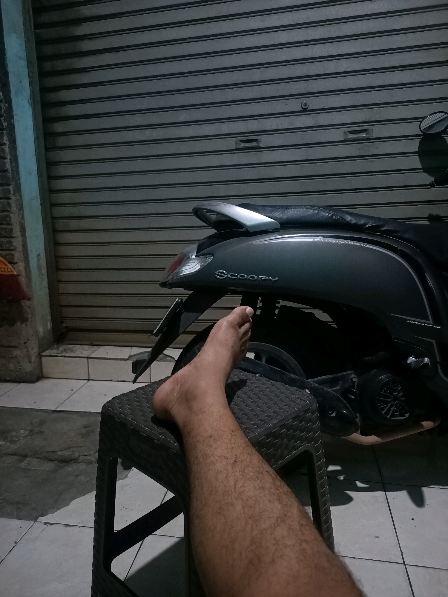 A person's leg is resting on a dark, textured plastic stool in front of a gray, corrugated metal roller door. The leg is tanned and hairy. To the right of the stool and leg, a dark gray scooter is parked. The scooter has a black seat and "Scoopy" is visible in silver lettering on its side. The background appears to be a wall and possibly a building exterior, with a tiled floor in front. The lighting suggests it is nighttime, with artificial light illuminating the scene. The image was captured in Telagaasih, Indonesia.