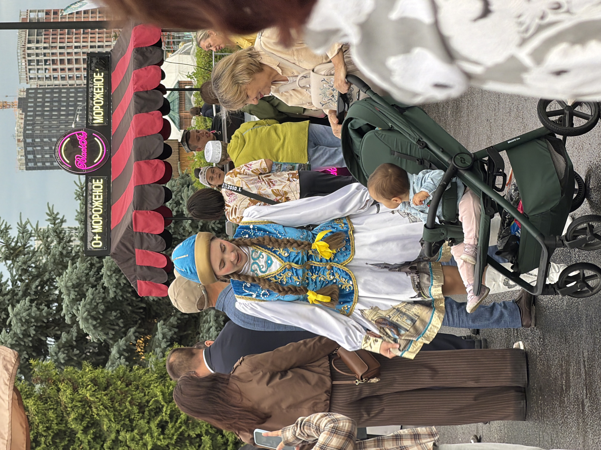 In an outdoor urban setting, possibly a market or festival, a young woman dressed in traditional clothing stands next to a stroller. The woman wears a blue and gold ornate vest over a white dress, with her hair in braids adorned with yellow ribbons. She holds a scarf patterned with buildings and gold trim. A baby sits in the dark green stroller, wearing a denim jacket and pink pants. A woman with blonde hair, wearing a light-colored shirt, is seen pushing the stroller. In the background, a kiosk with a red and black awning displays "МОРОЖЕНОЕ" (ice cream) in Cyrillic lettering, with a neon sign for "Black Ice" above it. Several other people are visible in the mid-ground and background, some wearing hats. The ground appears wet, suggesting recent rain. Tall trees are present on the left, and modern buildings under construction are visible in the distance.