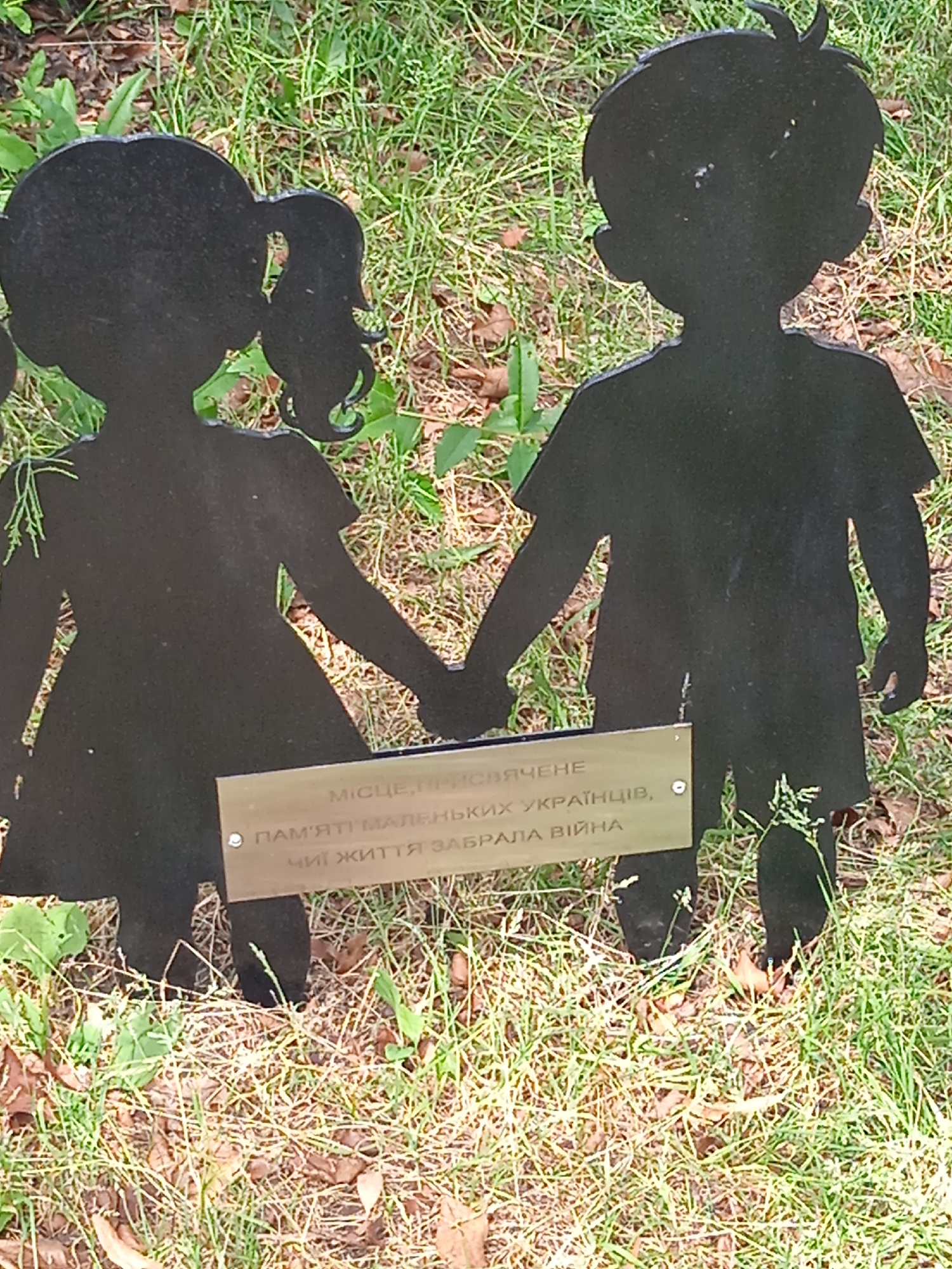 A silhouette of a boy and a girl holding hands stands in a grassy outdoor area. The silhouettes are black and appear to be made of metal. They are positioned in front of a small, metallic plaque with engraved text. The plaque has two lines of text. The first line reads "МІСЦЕ, ПРИСВЯЧЕНЕ". The second line reads "ПАМ'ЯТІ МАЛЕНЬКИХ УКРАЇНЦІВ, ЧИЇ ЖИТТЯ ЗАБРАЛА ВІЙНА". The background consists of green grass and some fallen leaves.
