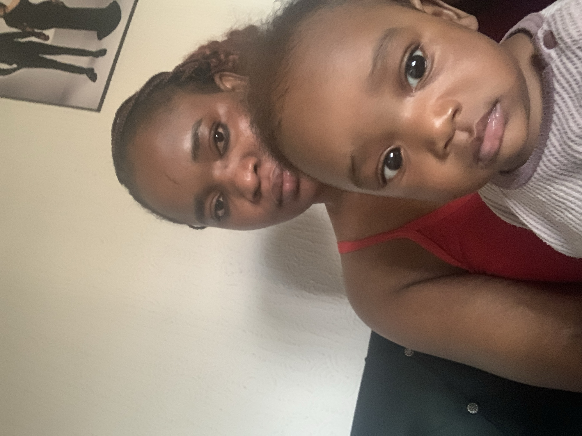 A close-up photograph depicts an adult female and a young child, possibly a baby. Both individuals have dark skin tones. The adult female has braided hair and is wearing a red tank top. The child is wearing a striped garment with a purple trim. They are positioned close together, appearing to be taking a selfie. A framed picture is visible on the wall behind them. The location is estimated to be Barakin Akawo, Nigeria.