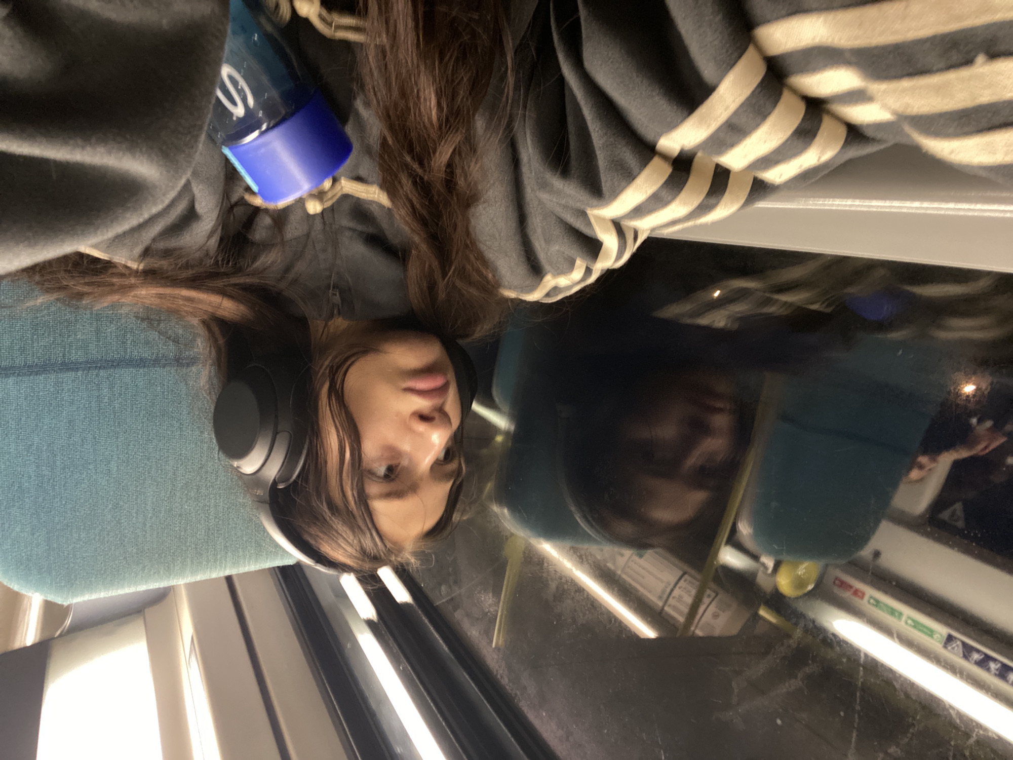 A young woman with brown hair wearing headphones is resting her head against a window on public transportation. She is wearing a dark-colored hoodie with white stripes on the sleeves. Next to her is a blue water bottle. The reflection in the window suggests she is on a train in London, United Kingdom.