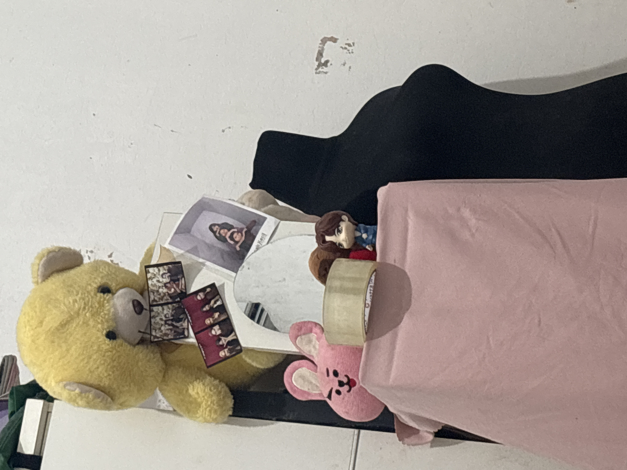 A stuffed yellow bear is positioned in the upper left portion of the image, holding several small photographs in its paws. To the right of the bear, a black turtleneck garment is displayed vertically against a white wall. In the foreground, a pink fabric is draped over an unseen object, with a roll of clear tape and two small figurines placed on top of it. The overall scene appears to be indoors, possibly in a domestic setting, within the city of North Jakarta, Indonesia.