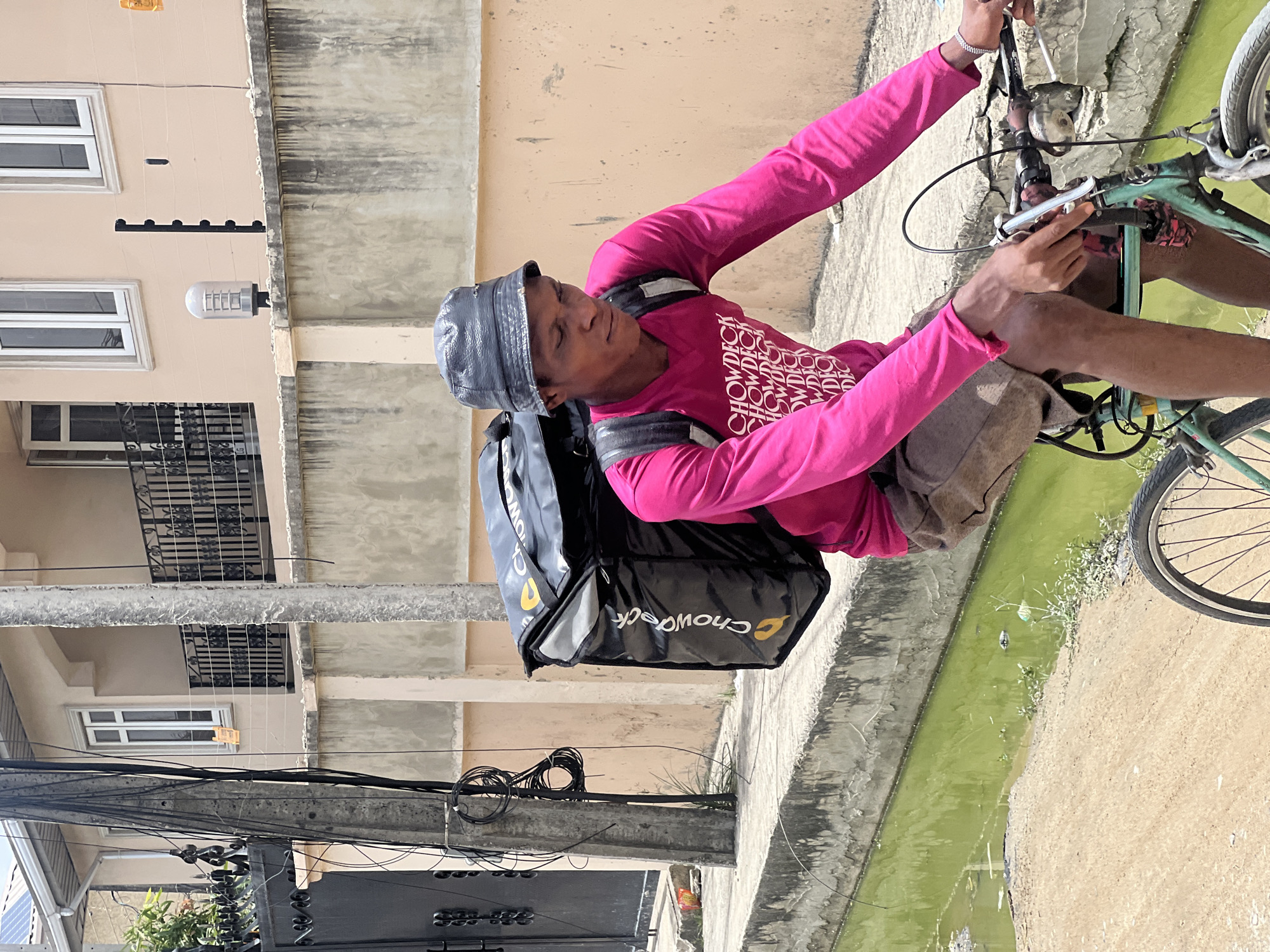 An outdoor scene in Igboefon, Nigeria, is captured on a bright, sunny day. The primary subject is a man, likely a delivery rider, seated on a bicycle. He is wearing a vibrant pink long-sleeved shirt with the word "CHOWDECK" printed repeatedly in white across the chest, grey shorts, and a grey bucket hat. A black delivery backpack with a prominent yellow "ChowDeck" logo is strapped to his back.
The man holds the handlebars of a green-framed bicycle, appearing to be in a moment of rest or preparation. His eyes are somewhat closed or squinting, possibly due to the strong sunlight. The background features a multi-story building with a light beige or cream facade, white-framed windows, and dark metal railings. An outdoor light fixture and security spikes are visible on the building's wall.
Below the building, a concrete drainage ditch filled with stagnant green water and some sparse vegetation runs parallel to a sandy path. On the left side of the frame, a utility pole is visible, heavily laden with a tangle of black electrical or communication cables. The clear skies and bright illumination suggest it is daytime, likely mid-morning or afternoon. The overall setting depicts a typical urban or peri-urban environment in Nigeria.