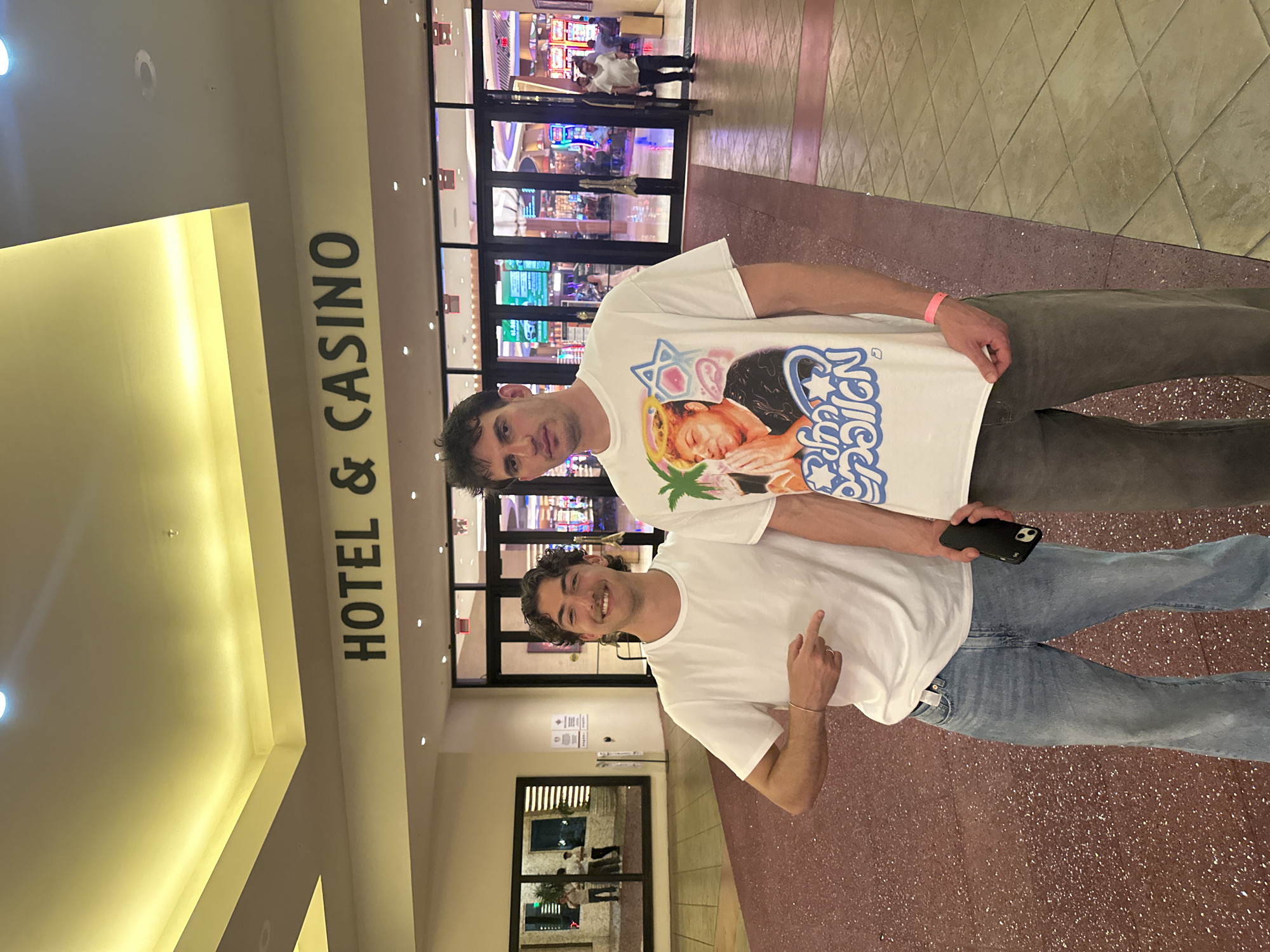 Two young adult males are posing in front of the entrance to a casino. Above them is the signage "HOTEL & CASINO". The casino entrance is visible in the background. The location is the Seminole Hard Rock Hotel & Casino in Hollywood, Florida, USA. The male closest to the camera is wearing a plain white t-shirt and blue jeans, pointing towards the other male, who is wearing a white graphic t-shirt and grey pants, and holding a mobile phone.