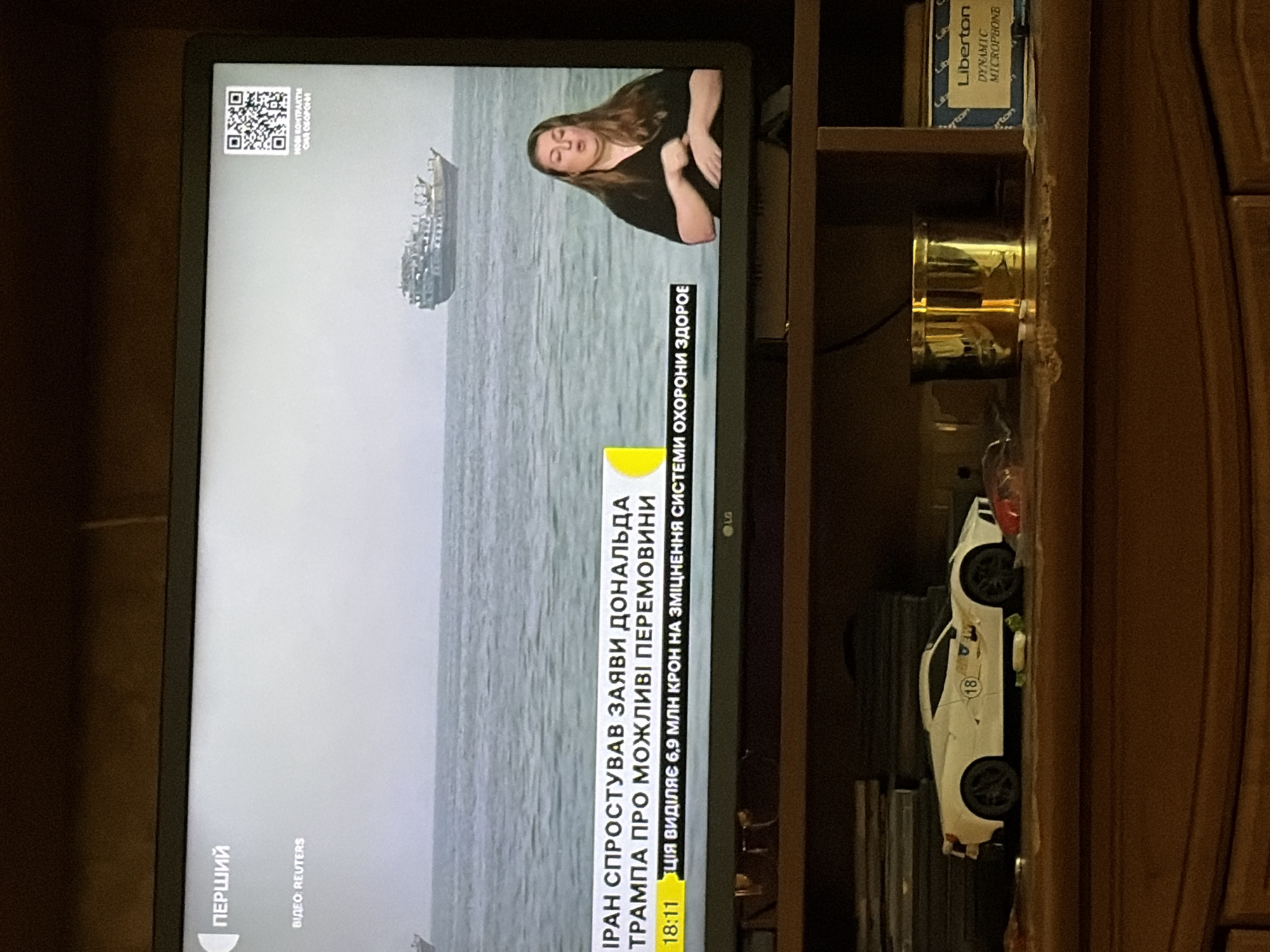 The image shows a television screen displaying a news broadcast. On the screen, there is a woman speaking, presumably a news anchor or interpreter, with a body of water and a ship in the background. Text overlays on the screen indicate "ПЕРШИЙ" (First) and "ВІДЕО: REUTERS" at the top left, and a news ticker at the bottom with information in Ukrainian. A QR code is present in the top right corner. Below the television, on a shelf, sits a white toy car and a gold-colored cylindrical object. To the right of these objects, a blue and white box with text "Liberton DYNAMIC MICROPHONE" is visible. The overall scene appears to be a domestic setting, possibly a living room.