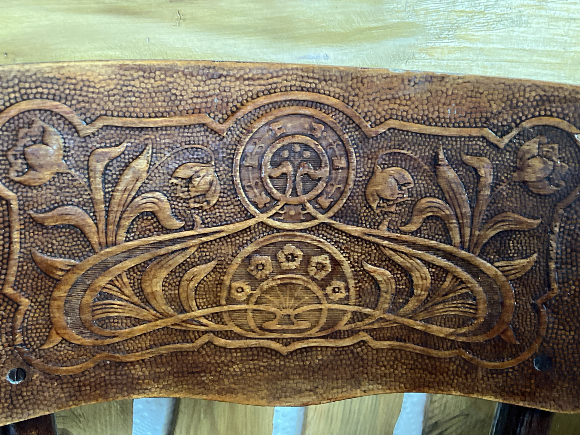 A close-up, eye-level, medium shot captures a detailed carved wooden backrest of a chair in Norviliškės, Lithuania. The wood is a dark, rich brown with a textured, stippled background. The carving features an ornate Art Nouveau style with flowing lines and botanical motifs. In the center, a circular medallion contains a stylized tree-like design. Flanking the medallion are floral elements with stylized buds and leaves. A lower section of the carving showcases a delicate arrangement of foliage and small flowers within a circular frame. The top edge of the backrest is smooth, while the bottom edge is slightly curved. The wood appears aged and shows subtle variations in color and texture, suggesting handcrafted quality.