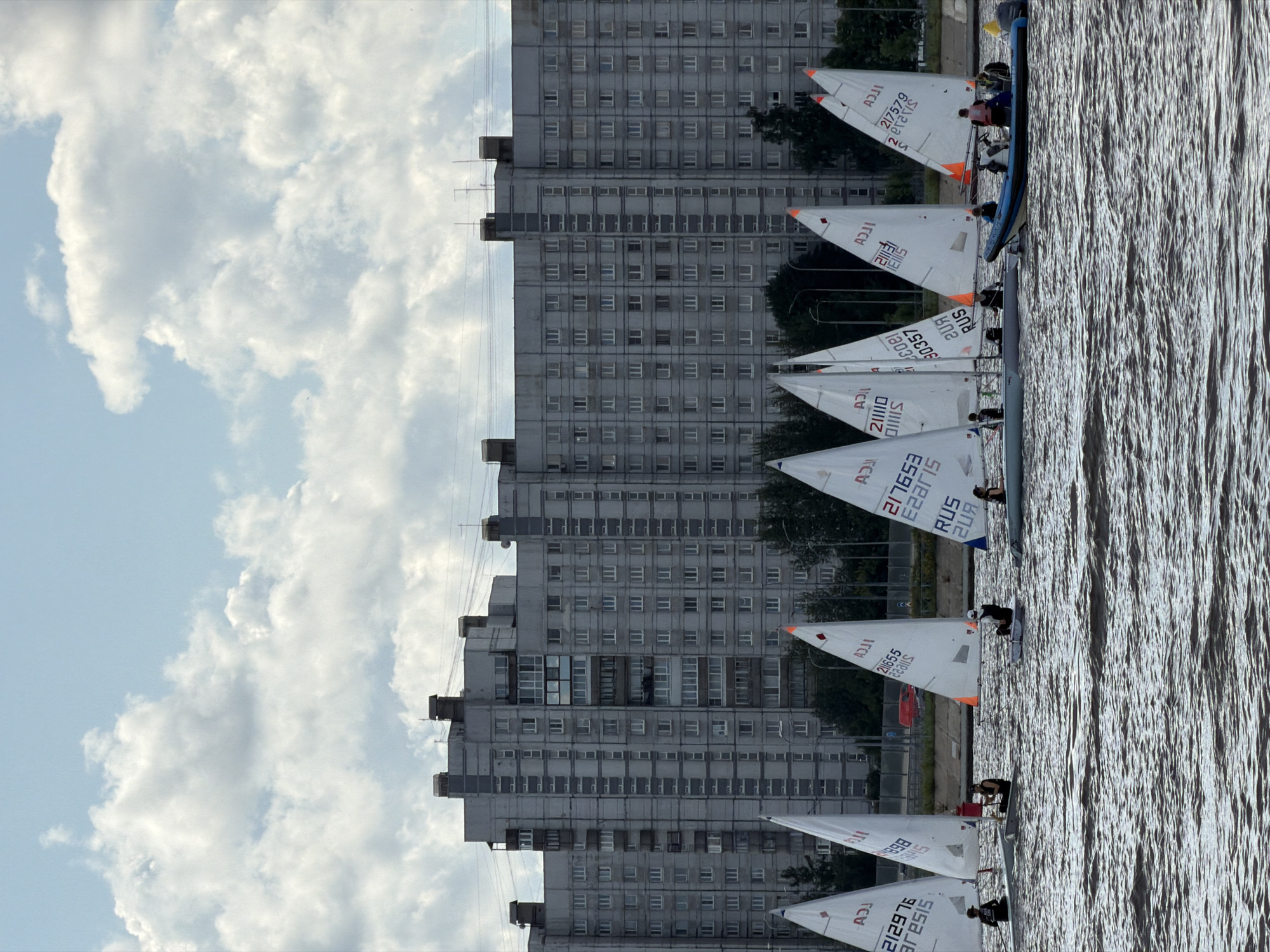 In Saint Petersburg, Russia, a fleet of small sailboats with white triangular sails is navigating choppy brown water. The sails bear black lettering and numbers, some of which are partially obscured. The boats are occupied by individuals dressed in dark clothing, some wearing life vests. In the background, a large, multi-story, gray apartment building with numerous windows dominates the skyline. Trees are visible in front of the building, and a blue and black inflatable boat with several people on board is to the right of the sailboats. The sky above is a mix of blue and white clouds. The scene appears to be a daytime event.