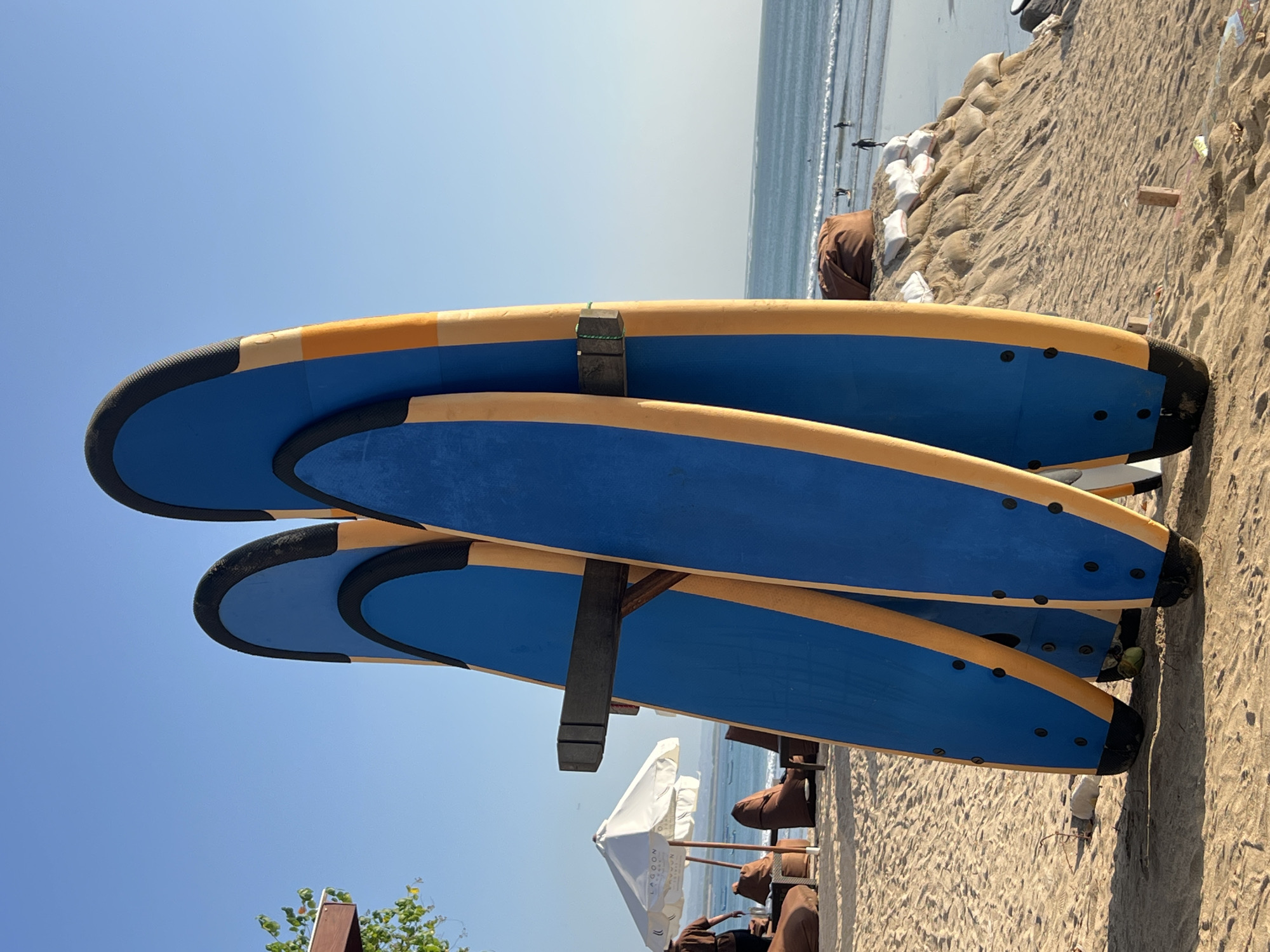 This image captures a sunny day on Kuta Beach, Indonesia, with a clear blue sky overhead. The foreground is dominated by a rack holding three blue and yellow surfboards, standing upright on the sandy beach. The surfboards are positioned at an angle, with the two on the left slightly behind the one on the right. A dark wooden bar is mounted on the surfboards, appearing to be part of the rack that holds them.
In the middle ground, a white umbrella stands open, casting a shadow on the sand. Nearby, several brown beanbag chairs are arranged, suggesting a relaxed seating area. A person, partially visible, is walking away from the viewer towards the umbrella.
Further in the background, the ocean stretches out to the horizon, with gentle waves rolling onto the shore. A few people can be seen in the water, some standing and others appearing to be surfing. The beach is lined with sand dunes, some of which are reinforced with sandbags. The overall atmosphere is one of a tranquil beach day.