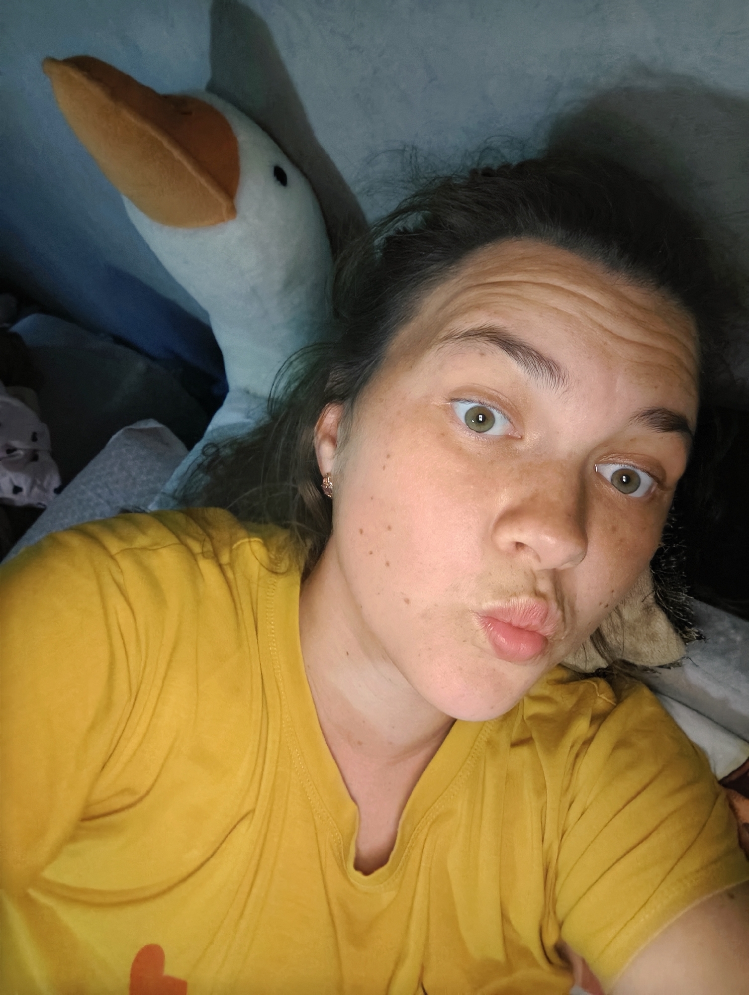 A person with light skin, freckles, and green eyes is posing for a selfie. Their hair is dark and pulled back messily. They are wearing a yellow T-shirt with a small orange heart on it. Their lips are pursed in a kiss. Behind them is a large, white, plush goose with an orange beak. The background is blurry and indistinct, appearing to be bedding or a cushioned surface. The location is Chaikovsky, Russia.