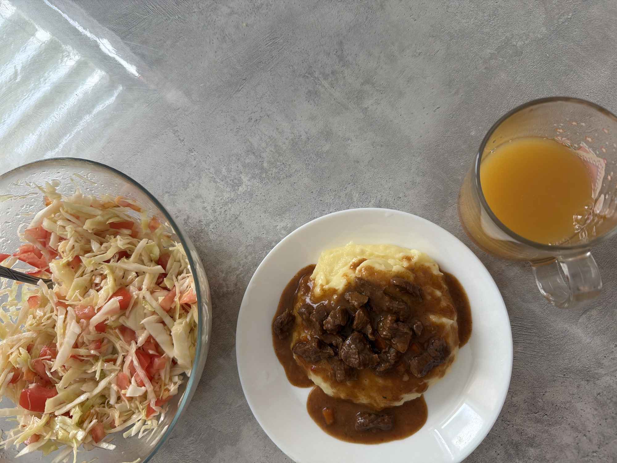 The image shows a meal setting on a gray surface. On the left, there is a clear glass bowl containing a salad of shredded cabbage and chopped tomatoes with a serving utensil inside. To the right is a white plate with mashed potatoes topped with pieces of meat in gravy. Next to the plate is a clear glass mug filled with orange juice. The upper left of the image contains an area of white plastic. There is no visible evidence to confirm the specific location within the city of Chaikovsky, Russia.