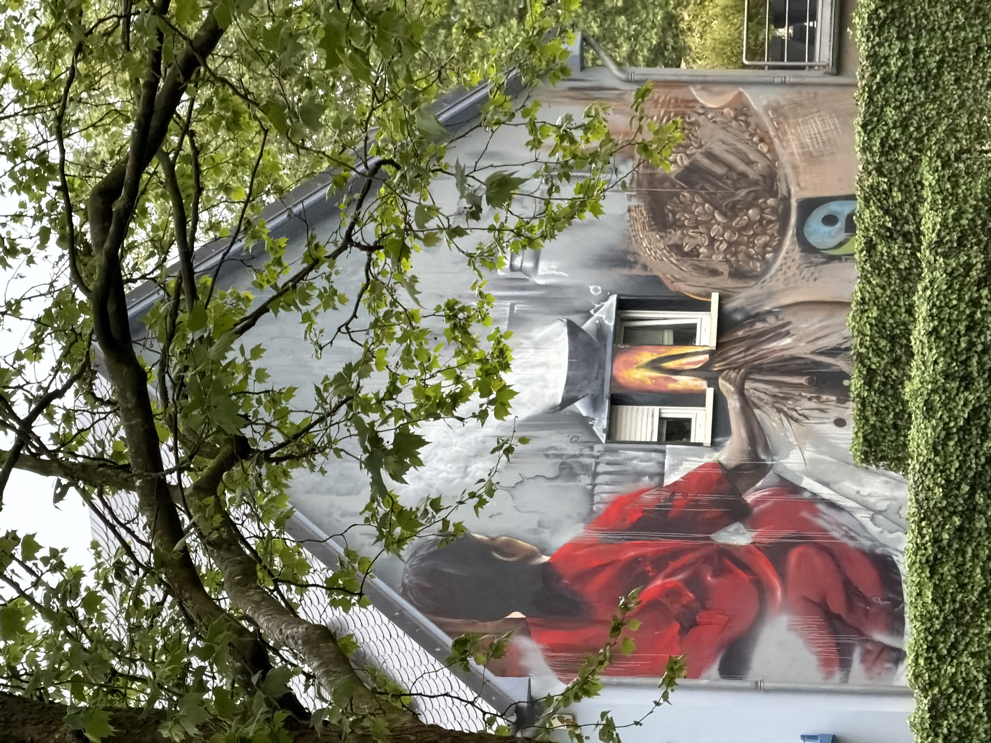 The image depicts a building with a large mural on its side, likely in Esch-sur-Alzette, Luxembourg. The mural features a person or figure dressed in red, appearing to lie down, and above them, an abstract image resembling a basket or container with a blue eye. The building has a grey roof and is partially obscured by a tree with green leaves in the foreground. A vertical strip of dense green foliage is visible on the right side of the building.