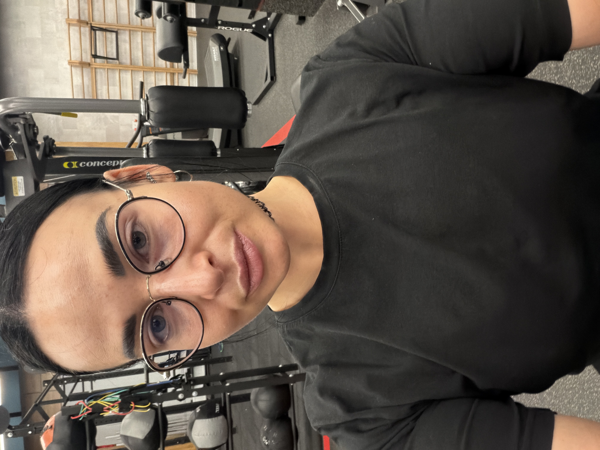 A woman with dark hair and glasses is in a gym. The background includes gym equipment such as a Concept2 rower, Rogue Fitness equipment, and medicine balls. The woman is wearing a black shirt. The location is likely indoors and no information indicates it is in St. Petersburg.