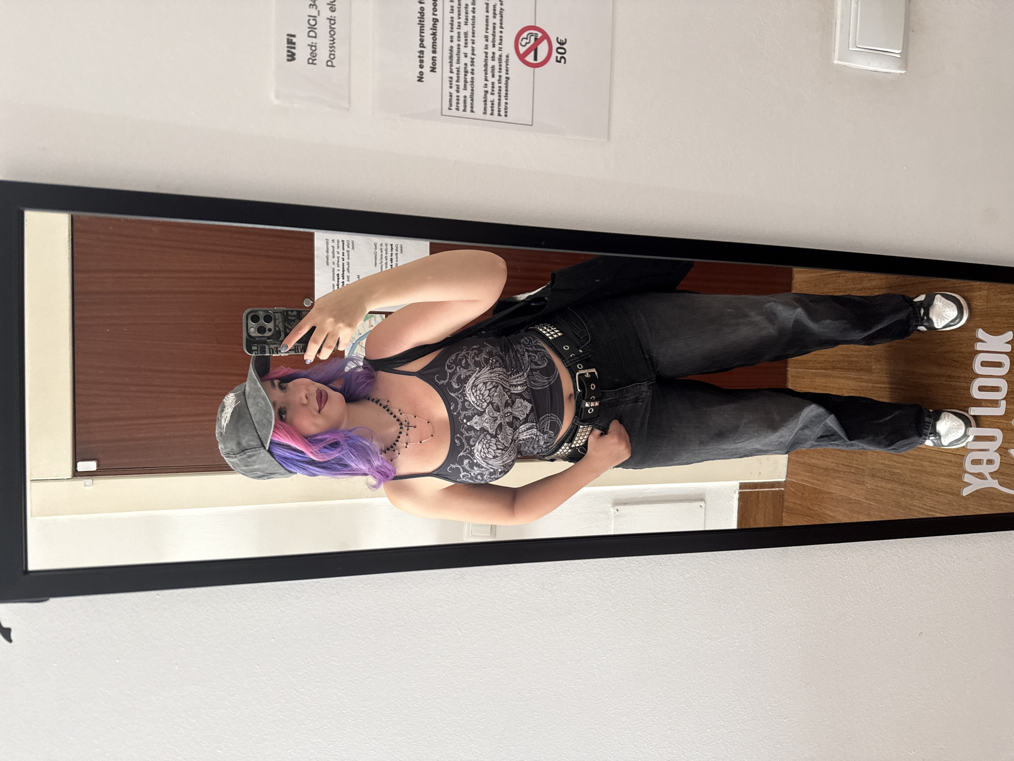 A woman with purple hair and a gray hat is posing in a full-length mirror. She is wearing a black tank top with a paisley pattern, black jeans with a studded belt, and black and white sneakers. She is holding a smartphone in her left hand, taking a selfie. A black blazer is slung over her shoulder. The mirror is mounted on a wall. Above the mirror are three white signs. The left sign has "WIFI" and network information. The middle sign states, "No está permitido fumar Non smoking room" and includes a fine of 50€. The sign on the right appears to be a light switch. The floor is wood-look laminate. The location is a building in Madrid, Spain, given the language on the signage.