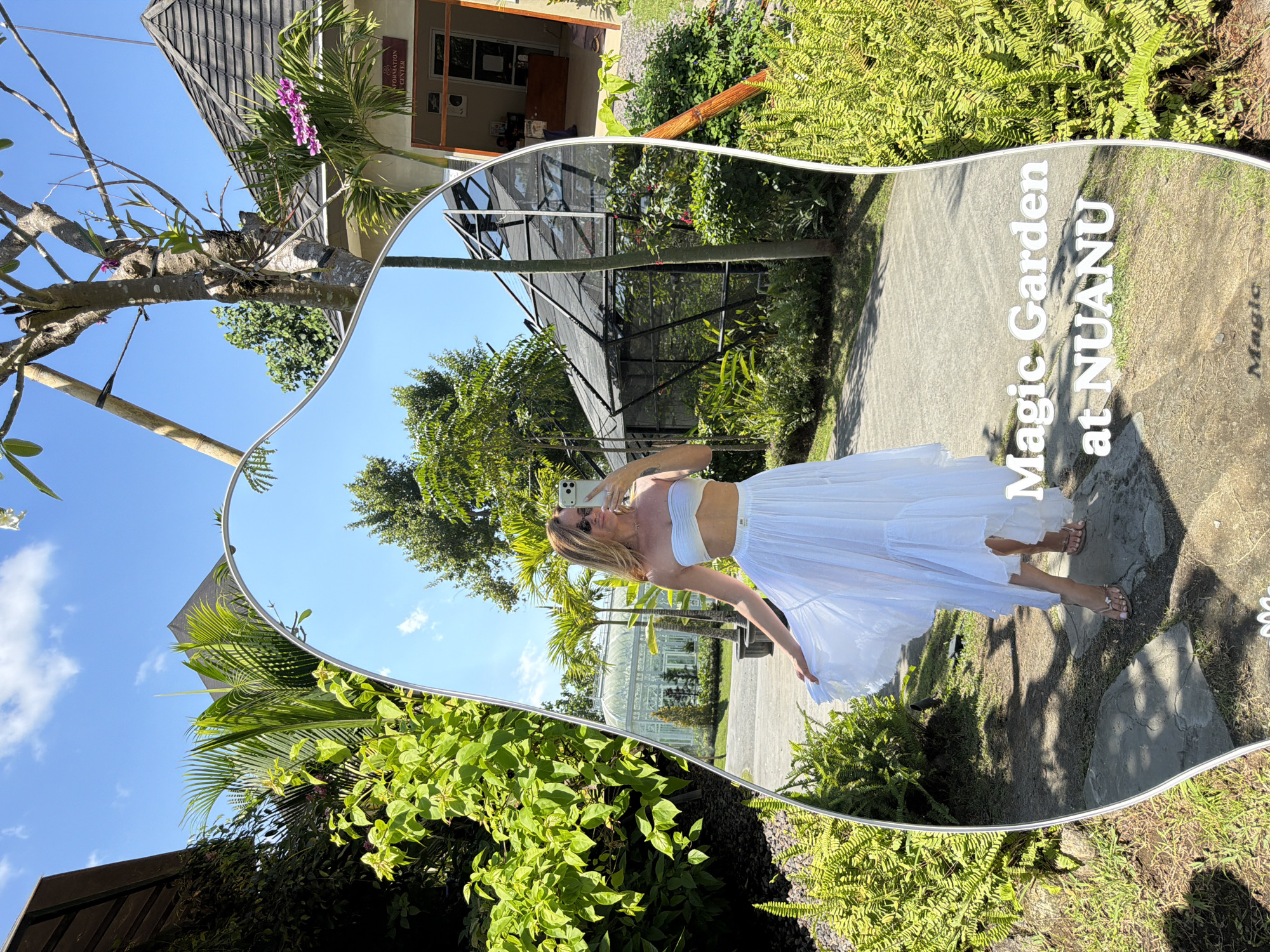 This is a medium shot taken in a garden with a large, irregular-shaped mirror as the central subject. The mirror reflects a woman in a white strapless bikini top and a flowing white skirt, holding a smartphone to take a selfie. She is wearing sunglasses and sandals. The background reflected in the mirror shows palm trees, a greenhouse-like structure, and a building with an "Information Center" sign. Lush green vegetation, including ferns and other plants, surrounds the mirror. The sky is blue with scattered clouds. Text overlaid on the mirror reads "Magic Garden at NUANU". The location is not Kediri, Indonesia.