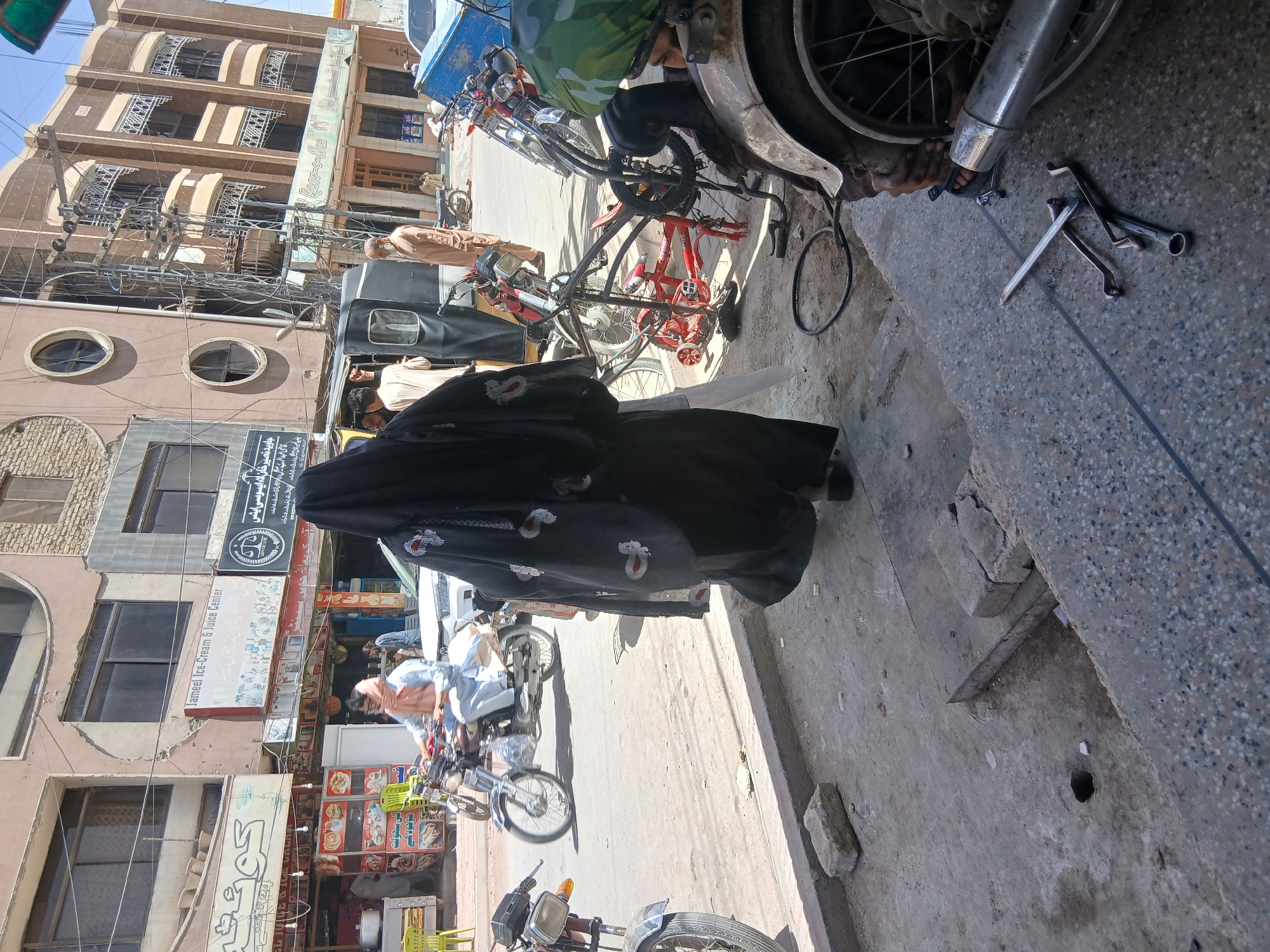 This image shows a street scene in Quetta City Tehsil, Pakistan. A woman wearing a black burqa with floral details is walking along the sidewalk, partially obscuring the view. Next to her, a mechanic is working on a motorcycle, with tools laid out on the ground nearby. Several other motorcycles are parked along the street. Buildings line the street, some with businesses such as Jameel Ice-Cream & Juice Center. Overhead power lines are visible. The environment appears urban and somewhat busy.