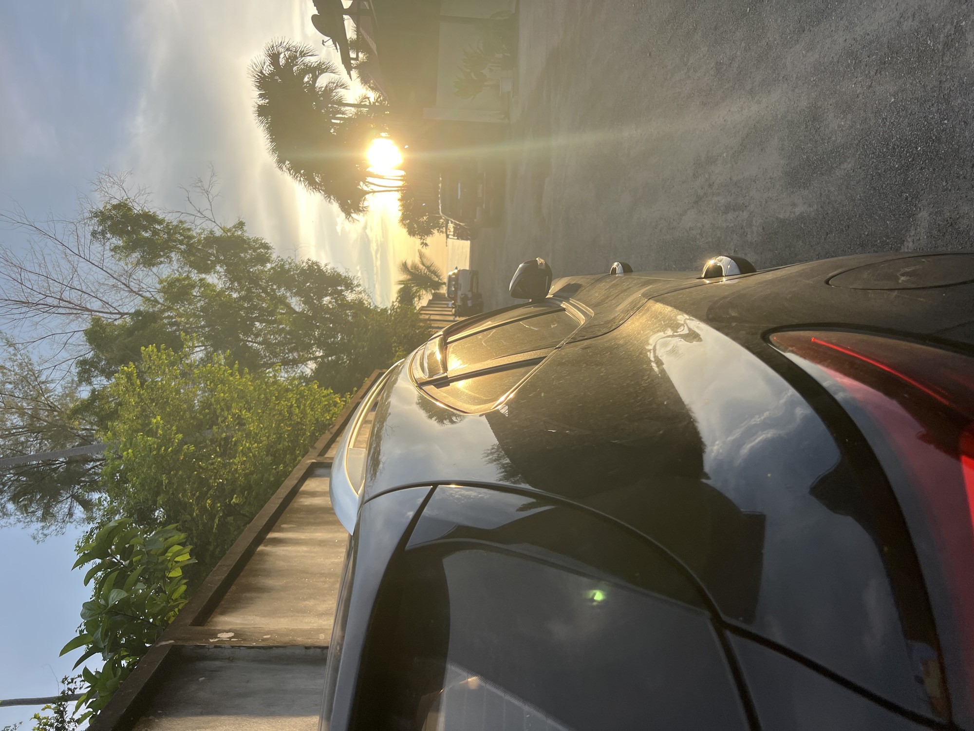 The image shows a dark-colored SUV parked on a gravel road. The sun is setting, casting a warm golden light across the scene. Lush green trees and palm trees line the background, with a concrete wall to the left. In the distance, other vehicles are visible, along with buildings and a glimpse of the ocean. The overall impression is one of a tropical setting at dusk.