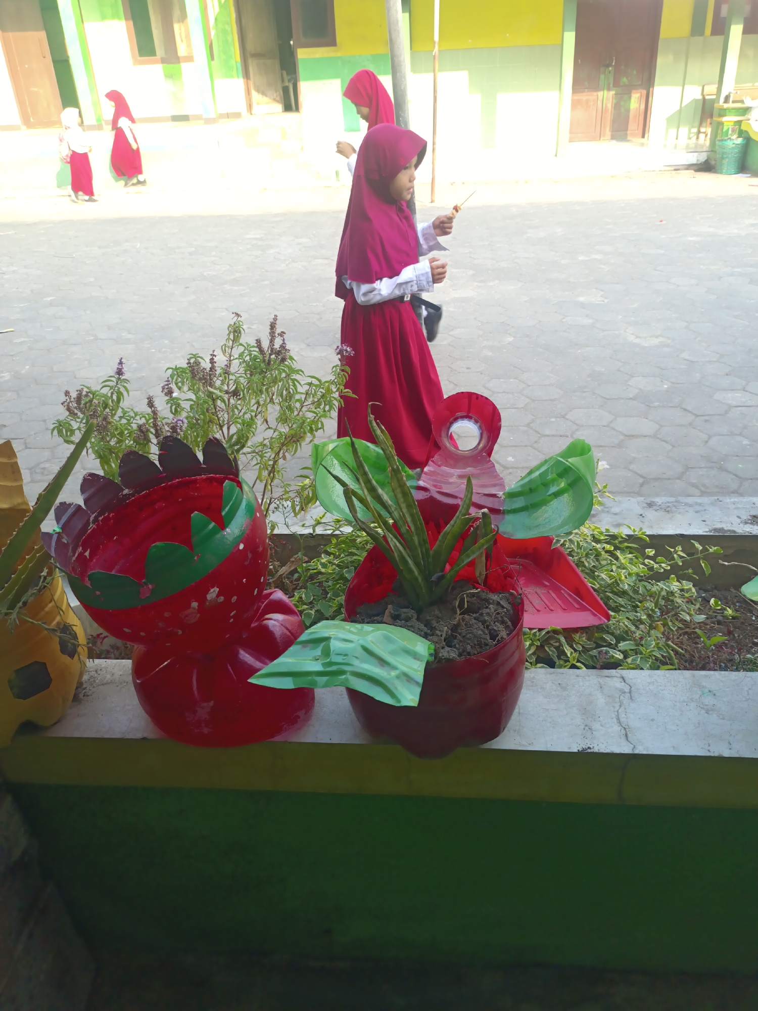 In Sarimulyo, Indonesia, on a paved walkway in front of a building with green and yellow accents, a young girl wearing a magenta hijab and a maroon dress stands holding a small stick with food on it. To her left, two other children, one in a white top and pink skirt and another in a pink hijab and skirt, walk away from the camera. In the foreground, creative planters made from repurposed red plastic bottles, shaped like a strawberry and a flower, hold plants. A tall, thin plant with sword-like leaves grows in the flower-shaped planter, while the strawberry-shaped planter is empty, its cut top revealing a dark interior. Green leaves and vines surround the planters, and a yellow planter is partially visible to the left. The scene is illuminated by bright sunlight, casting shadows on the ground.