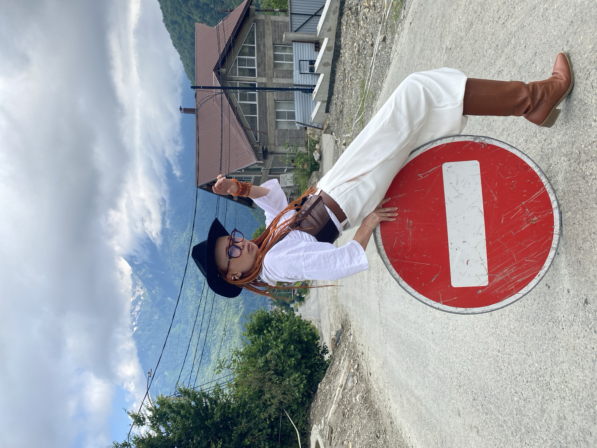 A woman with dreadlocks styled in braids, wearing a black hat, glasses, a white shirt, a brown corset, and white pants, sits on a round red and white "Do Not Enter" road sign on a paved road. She is leaning forward and to her right, with her right arm resting on the sign and her left arm raised. The road curves uphill and away from the viewer, with a forested mountainside on the left and a two-story house with a brown roof on the right. Power lines are visible across the upper portion of the image, with cloudy skies above the mountains. The scene appears to be in a rural or mountainous area, consistent with the location of Krasnaya Polyana, Russia.