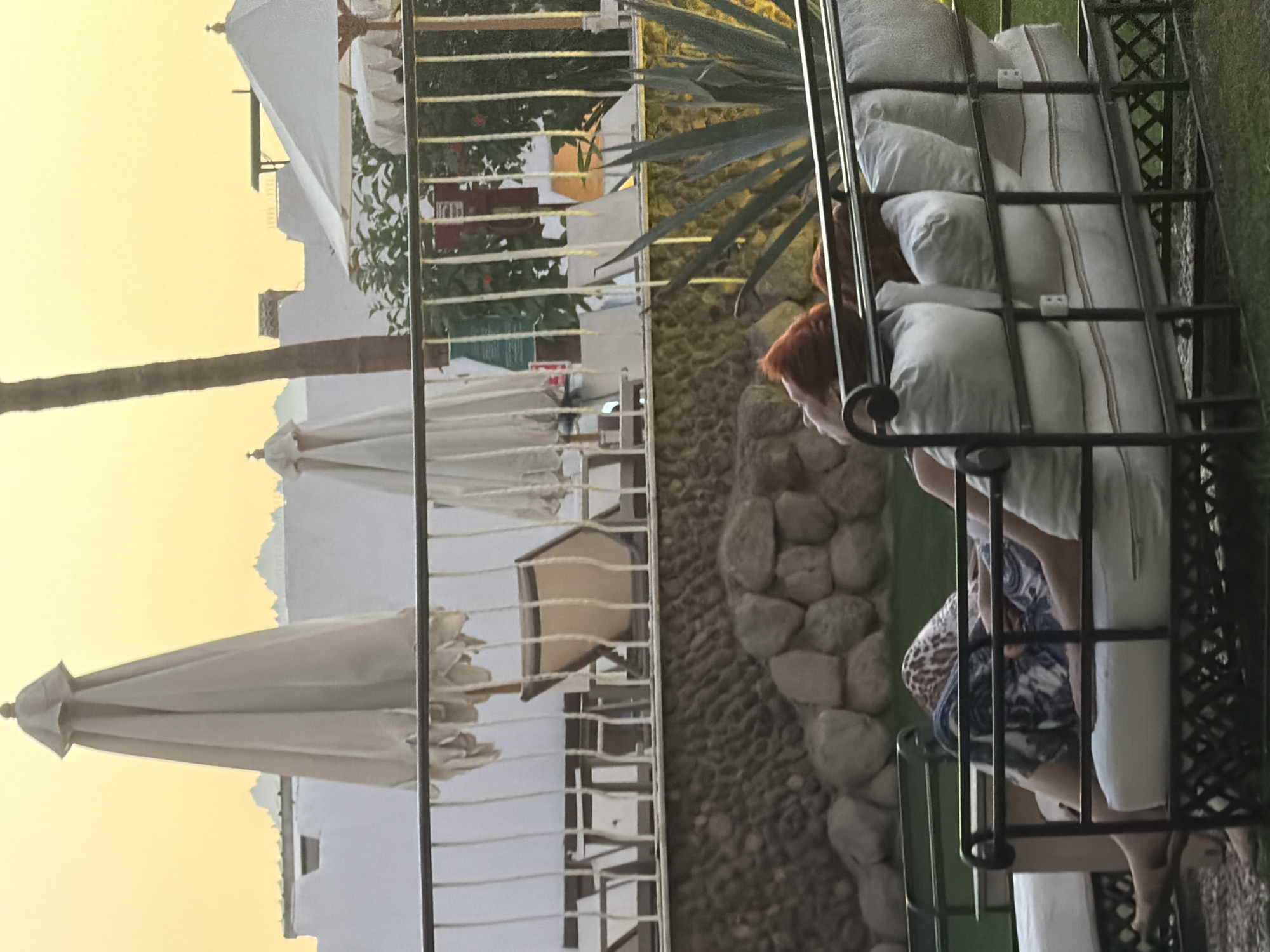 The image depicts an outdoor scene at what appears to be a resort or hotel in Sharm El Sheikh, Egypt. In the foreground, a woman with reddish-brown hair is reclined on a cushioned outdoor lounge bed. She is wearing patterned pants. The lounge bed is made of dark metal with white cushions. Behind the lounge bed, a metal railing separates the immediate area from a stone-walled terrace. On the terrace, there are several white patio umbrellas, some of which are closed. In the background, white buildings and palm trees are visible, and the sky has a warm, yellowish hue, suggesting either sunrise or sunset. A tall palm tree trunk is prominent on the right side of the image. The overall environment suggests a relaxed, possibly vacation-oriented setting.