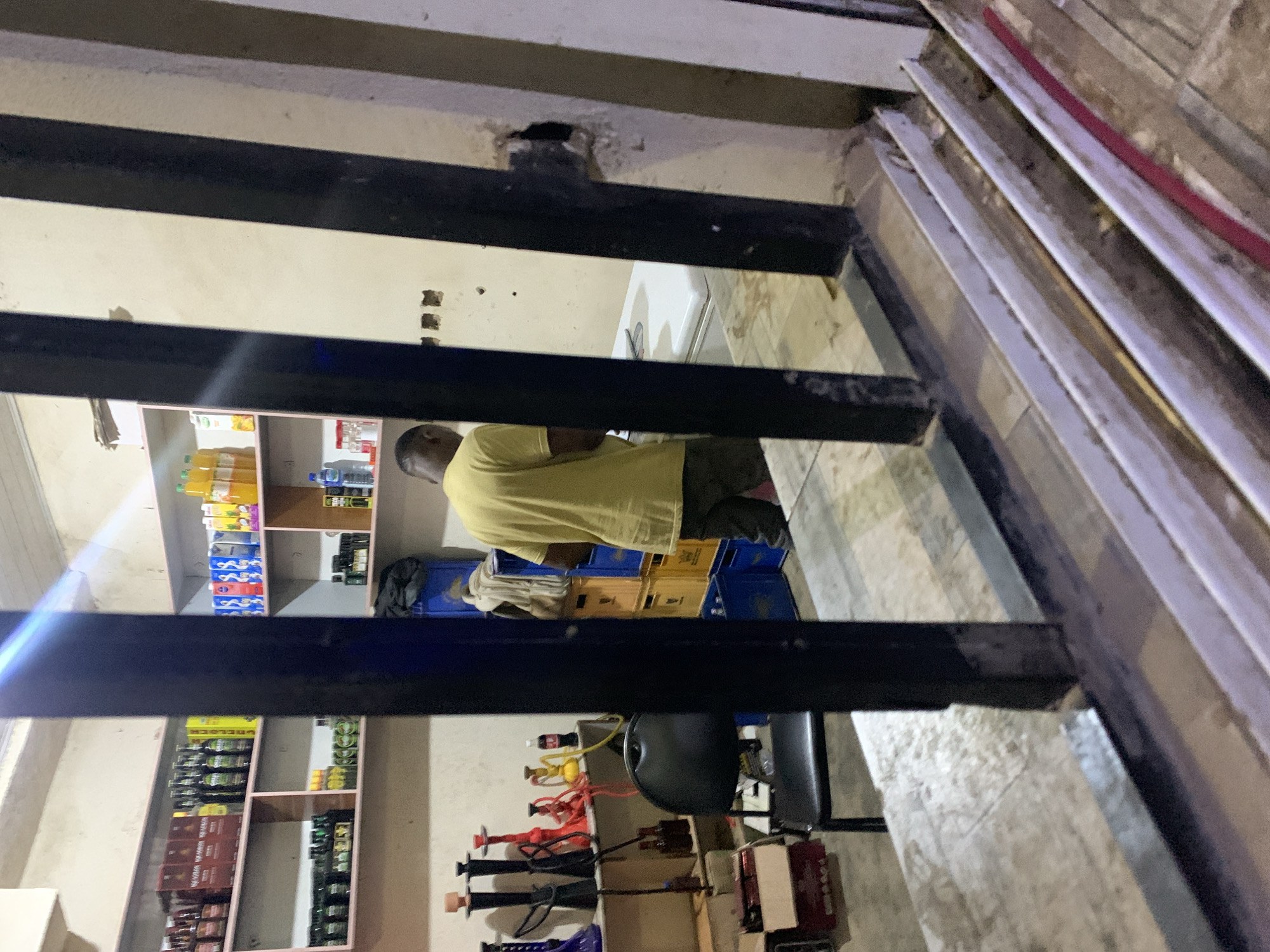 A person in a yellow shirt stands in a retail store in the New Stadium area of Nigeria, visible through the window bars. Shelves are stocked with drinks, liquor, and other items. Boxes and equipment sit on the floor.