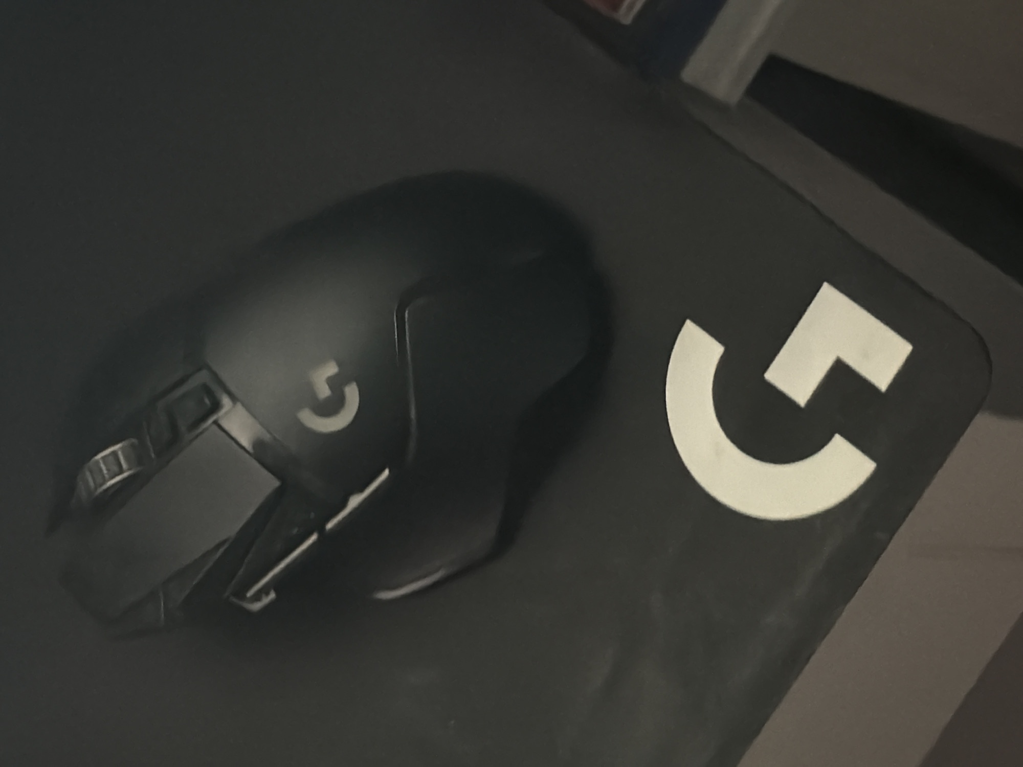 A black Logitech gaming mouse is positioned on a black mousepad. The mousepad features a large white "G" logo, representing the Logitech G brand. The scene is viewed from a high angle, with the mouse angled downwards and to the left. The lighting is dim, suggesting an indoor setting. There is no indication of the location being in Esch-sur-Alzette, Luxembourg, or any other specific city.