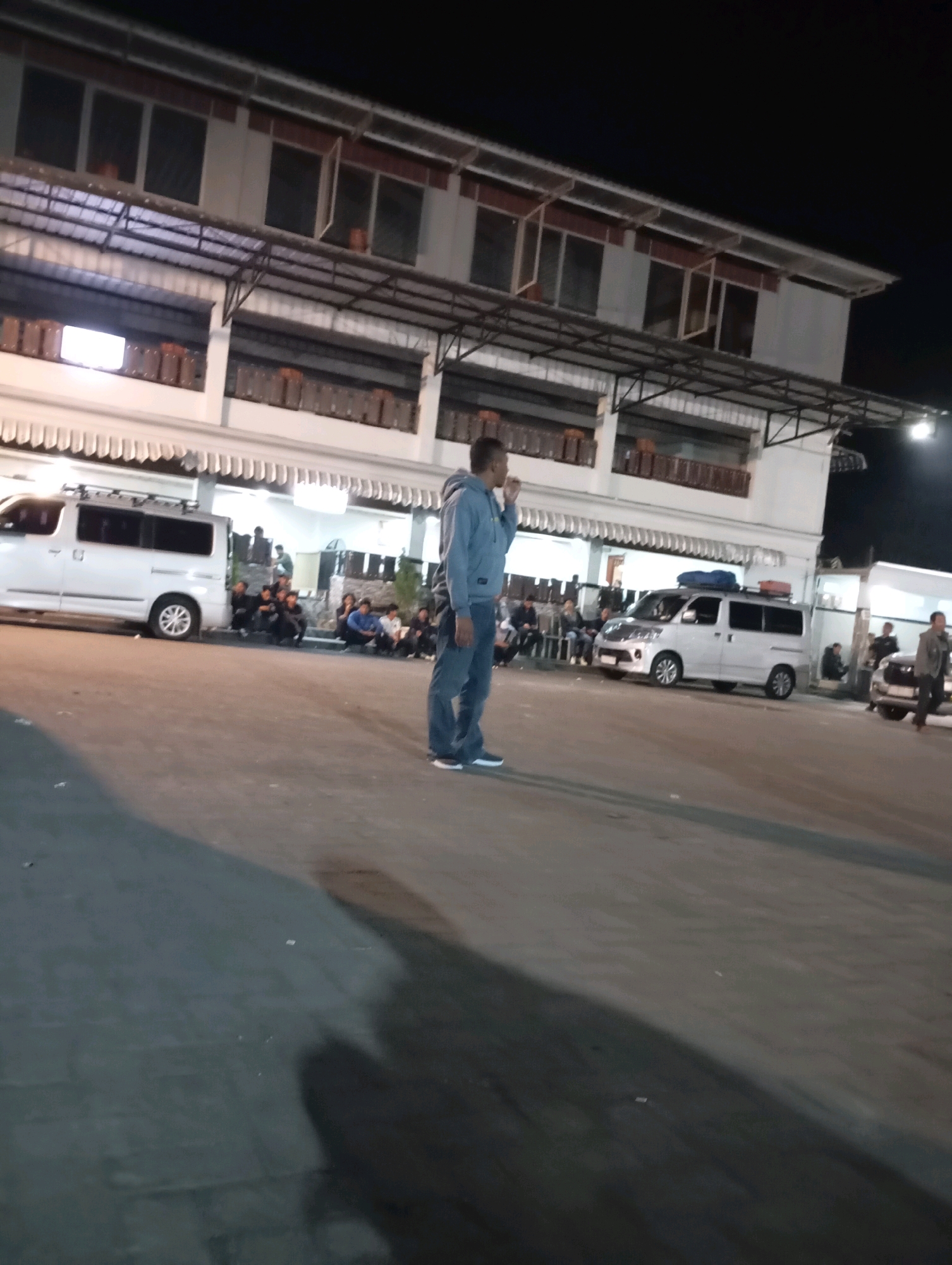 A man in a blue hoodie and jeans stands on asphalt, looking towards his right. Behind him, a white minivan is parked. In the background, a two-story building with multiple windows and awnings is visible, with more vans and a few people gathered near the building entrance. The scene appears to be an outdoor parking area at night, possibly near a market or commercial establishment in Purworejo, Indonesia.