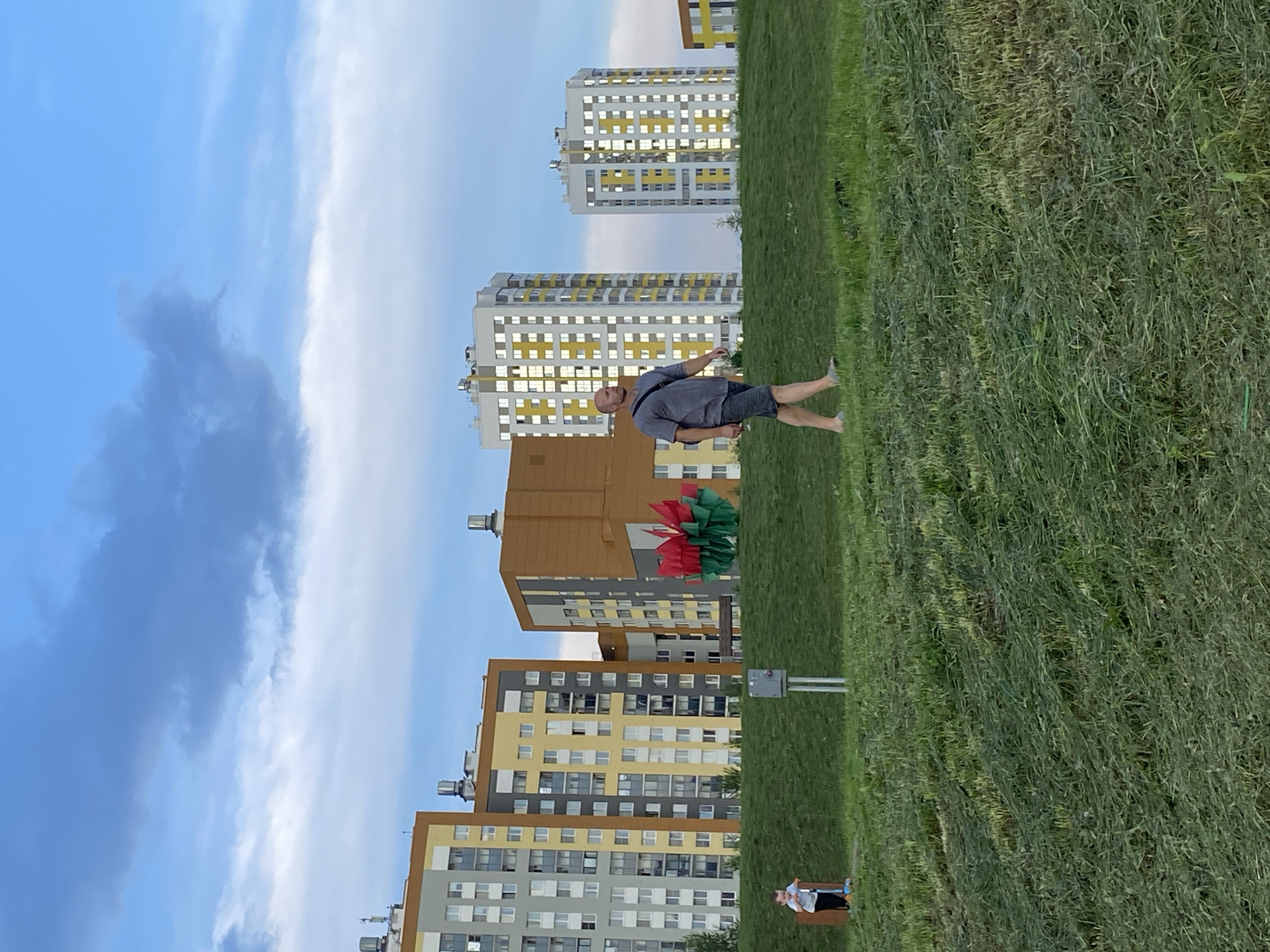 A man walks on a grassy hill in front of a row of modern apartment buildings in Yekaterinburg, Russia. The buildings are tall and multi-colored, with windows reflecting the sky. A child on a scooter is visible on the left side of the hill. In the center of the image, a large, red and green sculpture resembling a flower or plant is placed on the grass. The sky is blue with scattered clouds. The scene appears to be a park or recreational area within a residential complex.