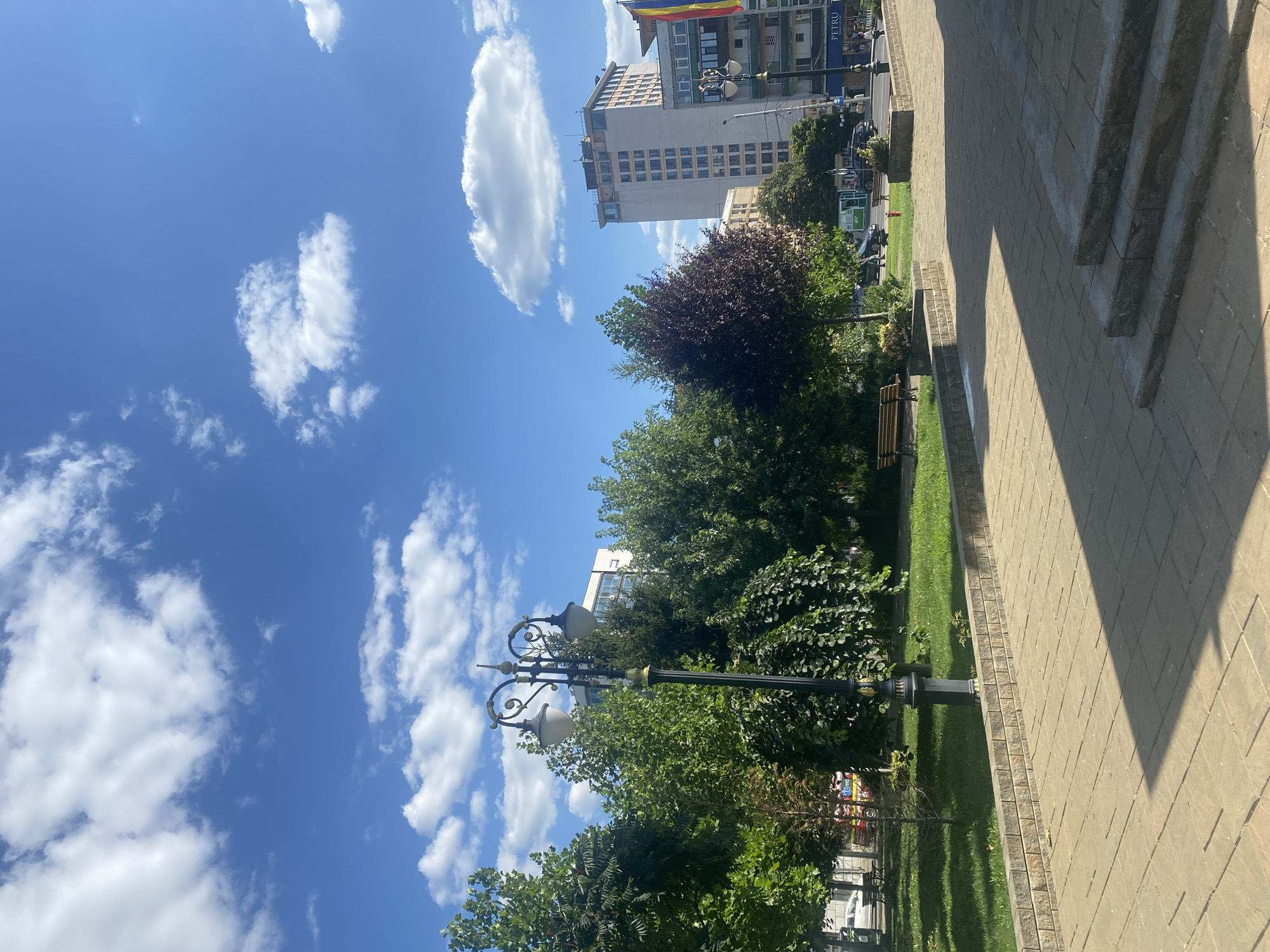 The image depicts a sunny day in Iași, Romania, likely in the city center, characterized by urban elements and lush greenery. In the foreground, a paved walkway with a shadowed area due to a nearby structure leads towards steps. A low brick wall borders a grassy area with several trees and a park bench. A ornate street lamp stands prominently in the middle ground. In the background, modern buildings, including a tall, light-colored structure with many windows, are visible, along with shops and a Romanian flag. The sky is a vibrant blue, dotted with scattered white clouds. No people are clearly visible or engaged in any specific action.