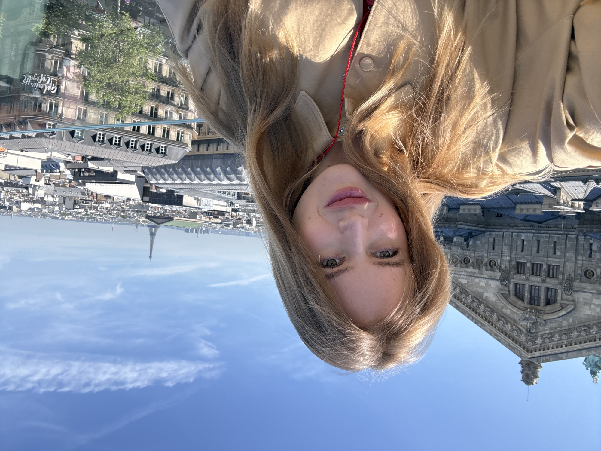 A woman with blonde hair wearing a tan jacket is facing the camera, with the city of Paris, France in the background. The Eiffel Tower can be seen in the distance. The sky is blue with some clouds. To the right, the architecture of a building is visible.