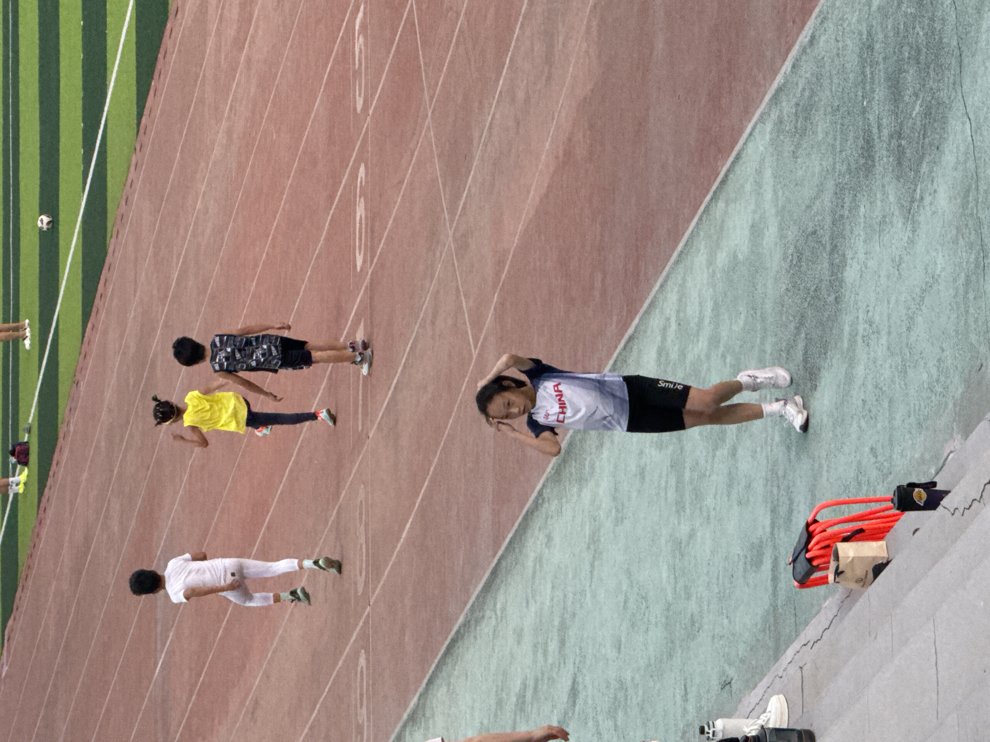 The image depicts a group of children on a running track, likely in a stadium located in Linghe, China. In the foreground, a young girl with dark hair is standing and touching her head, wearing a white and blue t-shirt with "CHINA" printed on it and black shorts. Behind her, further up the track, three other children are visible. One boy in a white t-shirt and white leggings is running with his right leg raised. To his right, a girl in a bright yellow sleeveless top and dark leggings is also in motion. Another boy, wearing a patterned sleeveless shirt and dark shorts, is walking further away. The running track is red with white lane markings. Beyond the track, a green field with striped patterns is visible, and a soccer ball is present on the field. On the left side of the image, a set of concrete stairs leads down to a green surface. Various objects are placed on these stairs, including white sneakers, a white water bottle, a brown paper bag, a black water bottle, and a bright orange structure with coiled black bands. The overall scene suggests an athletic activity or training session for young individuals.