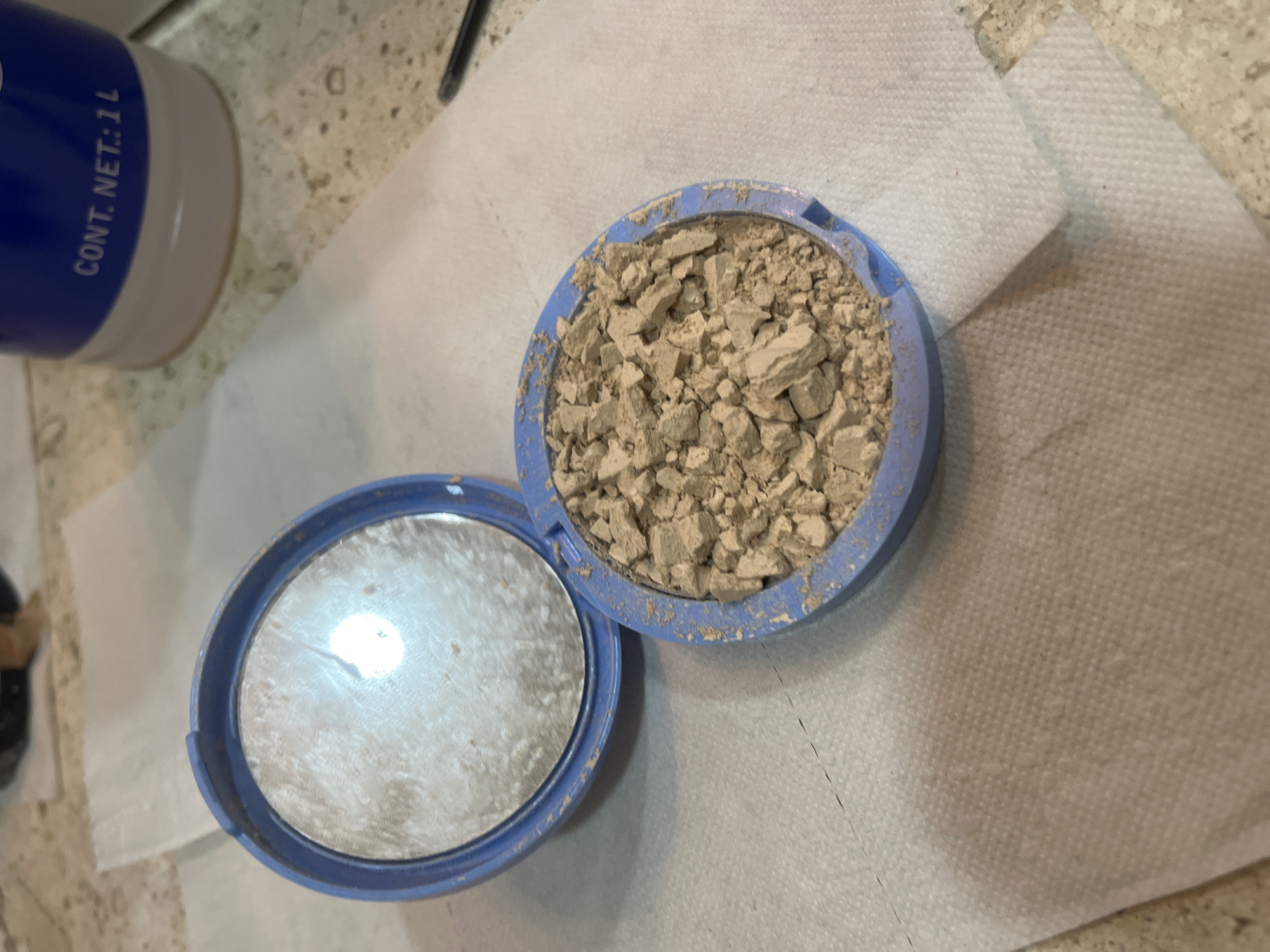 The image shows an open, blue compact of face powder sitting on a white paper towel on a countertop. The face powder is broken into chunks. The lid of the compact, containing a mirror, is open. A blue and white bottle labeled "CONT. NET.: 1L" is partially visible in the upper left corner. The countertop is speckled. The scene is forensic because the powder has been broken into chunks and has a gritty texture.
There are no people visible in the image. No location can be pinpointed.