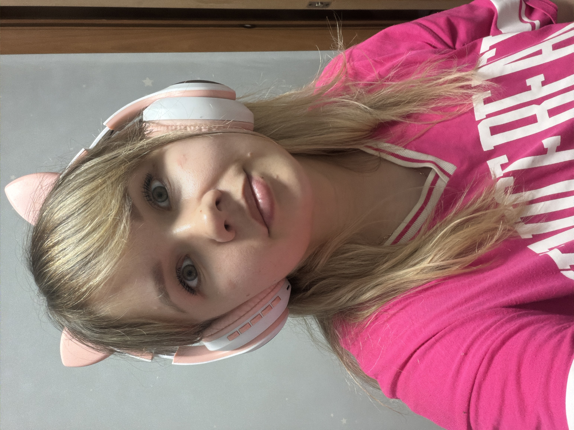 A young person with blonde hair and green eyes is wearing pink and white headphones with cat ears. They are wearing a pink shirt with white writing. The background is a light gray wall with small white stars. A dark wooden trim is visible at the top of the frame. The location and city are unknown.