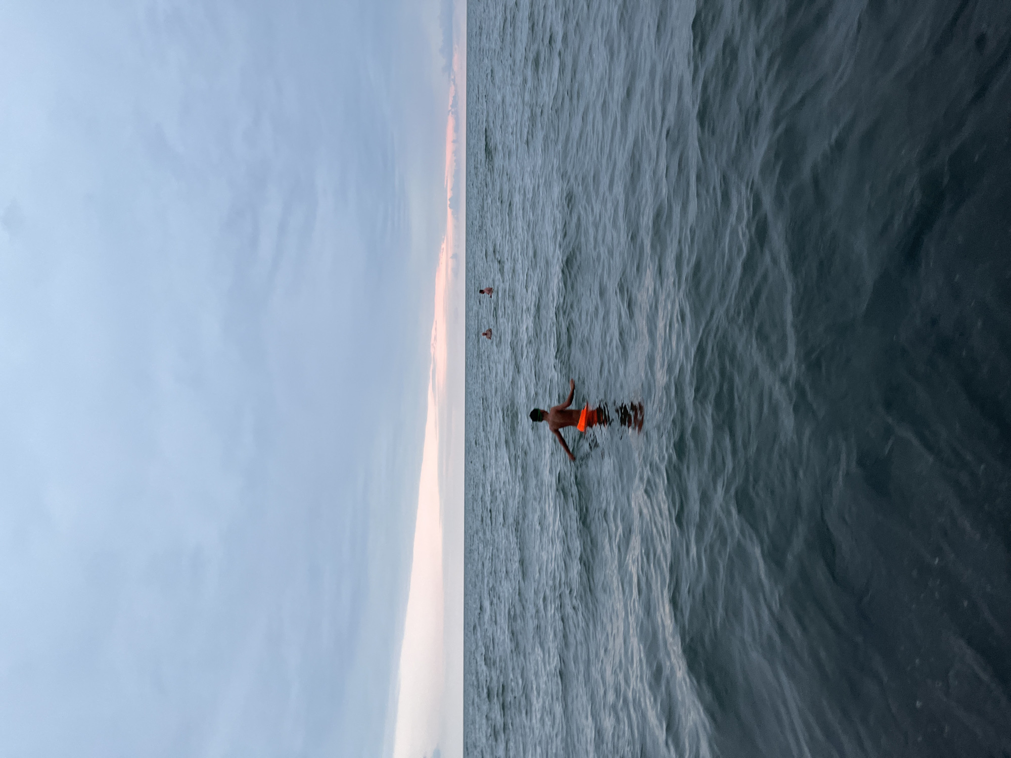 A view of the Black Sea from Ureki, Georgia, with three people swimming in the water. The sky is overcast with pastel hues near the horizon, suggesting either dawn or dusk. The sea is a deep blue-grey with small waves. The closest person is a young male wearing orange swim trunks, facing away from the camera and treading water. Two other individuals are visible further in the distance, also in the water. The overall scene conveys a sense of peaceful solitude.
