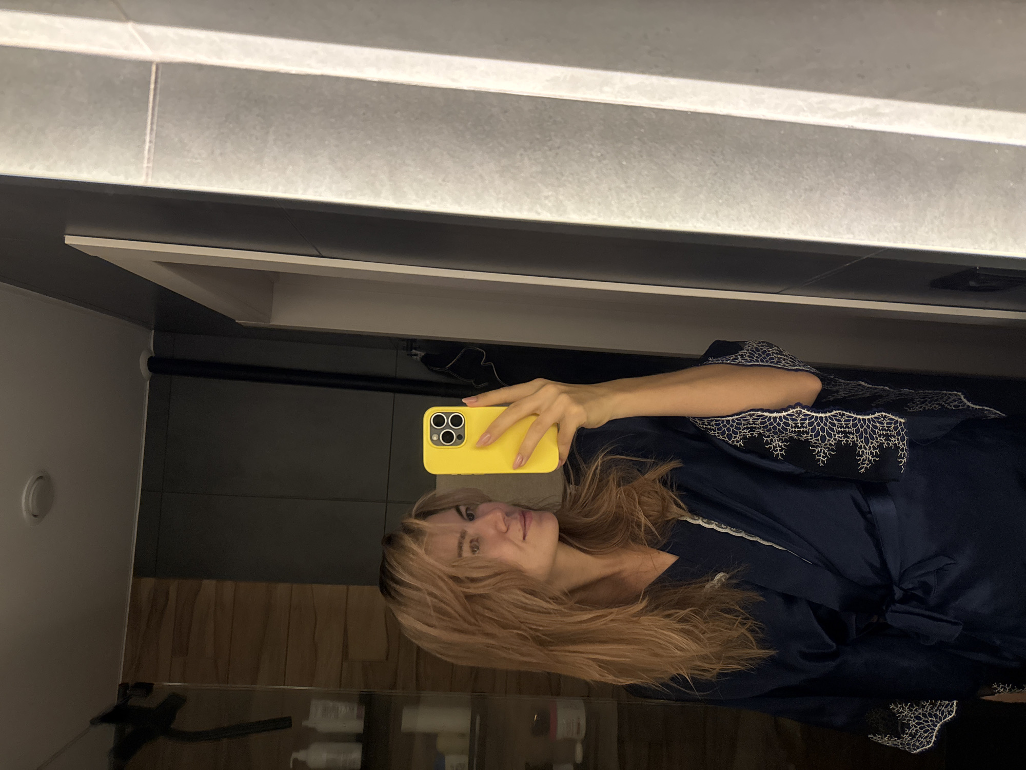 A woman with blond hair is taking a selfie in a bathroom. She is wearing a navy blue robe with white lace trim. She is holding a yellow smartphone. The bathroom has gray tiles and a black countertop. There are toiletries on the countertop. The location is Unknown, Unknown.